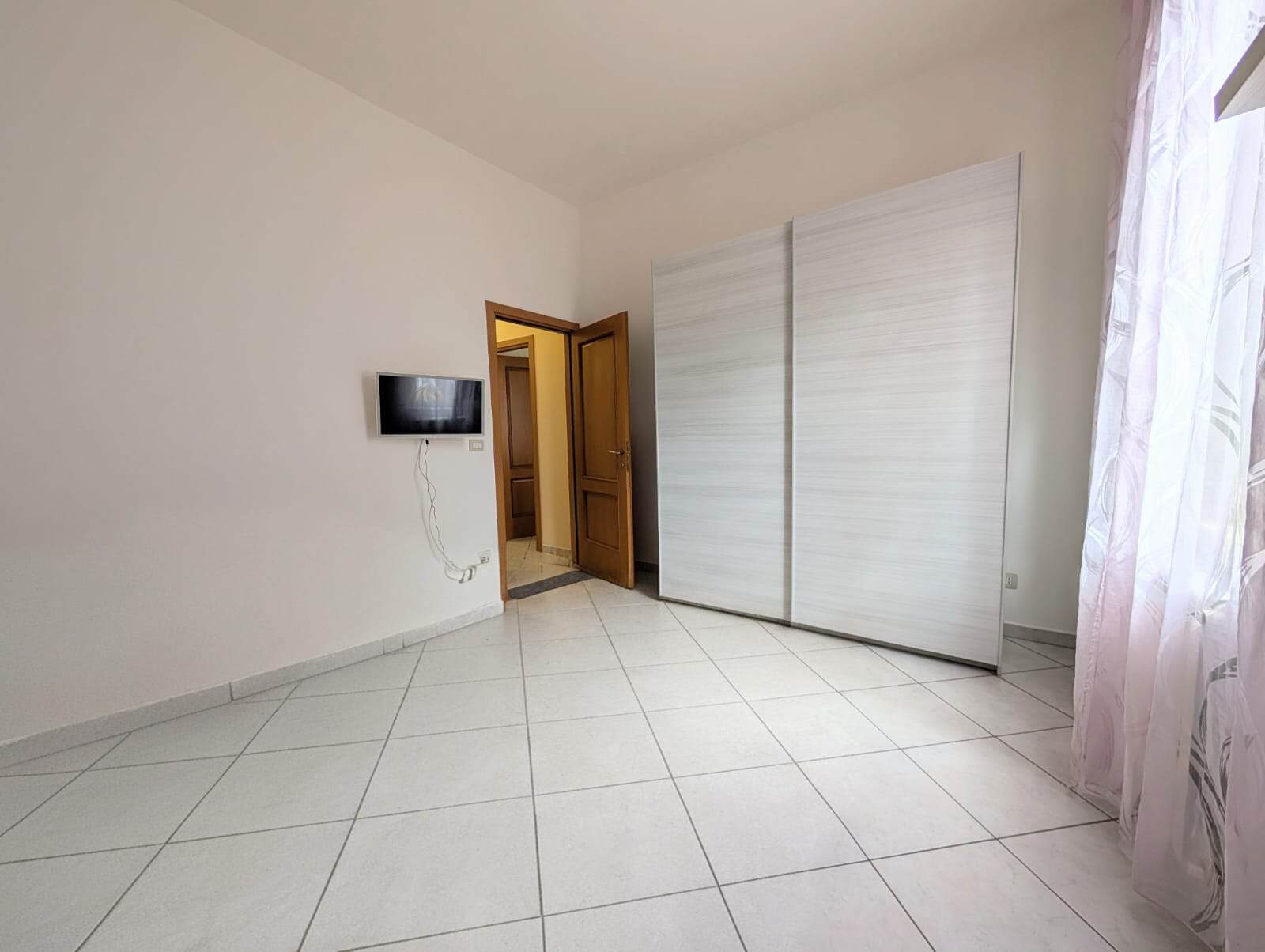 Camera da letto vuota con armadio bianco e finestra con tende leggere, ambiente luminoso e minimalista, ideale per affitti o acquisti immobiliari.