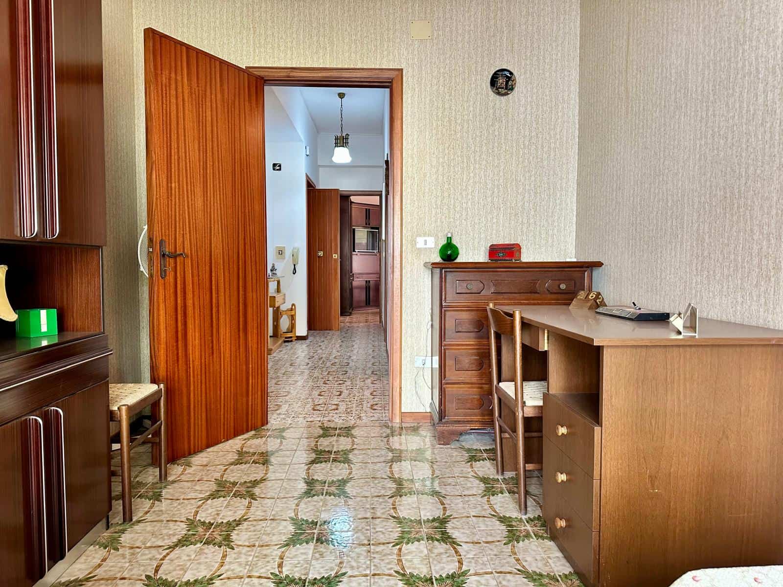 Porta aperta che conduce a un corridoio in un appartamento con arredamento in stile vintage, pavimento in piastrelle con motivo floreale, pareti beige e mobili in legno scuro.
