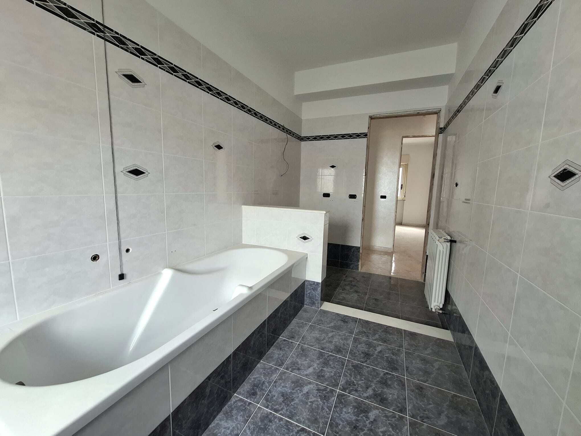 Bagno in fase di ristrutturazione con vasca da bagno moderna, pareti rivestite in piastrelle chiare e dettagli decorativi neri, spazio luminoso e funzionale.