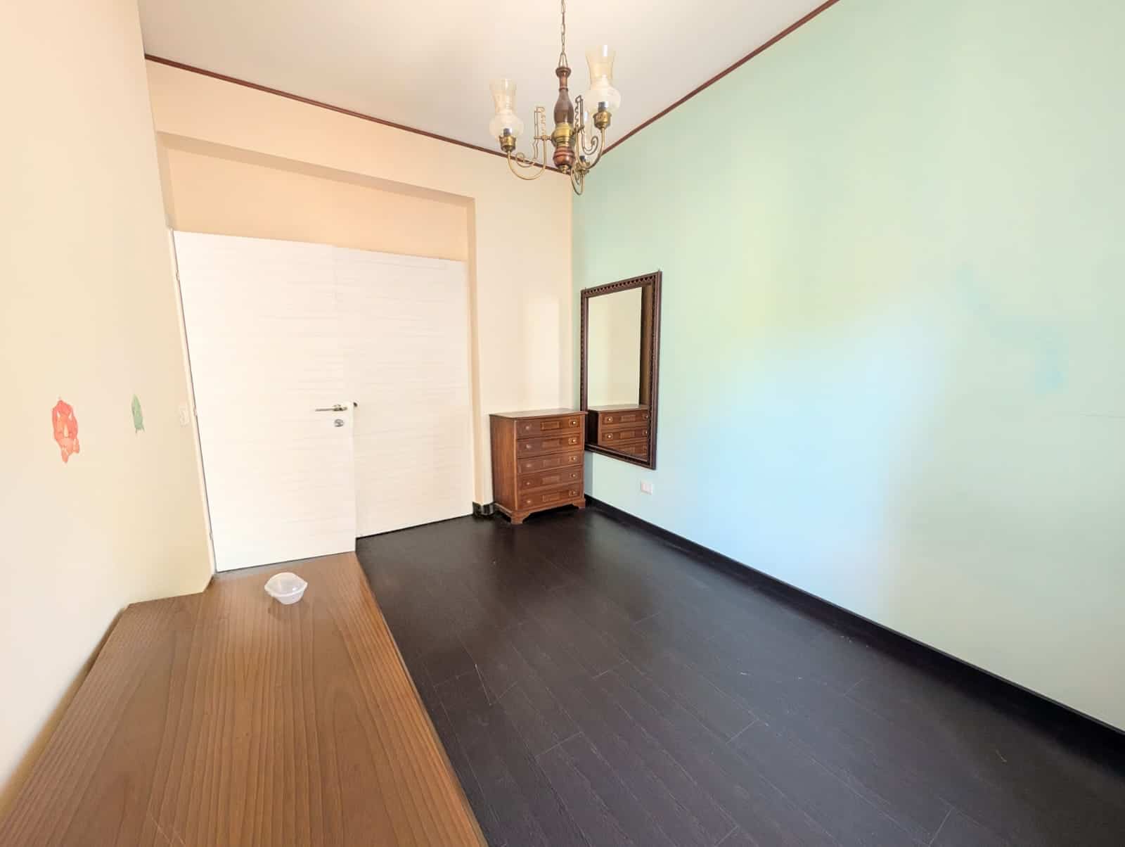 Lavagna di una stanza vuota con pareti colorate, pavimento in legno scuro e lampadario antico, arredamento minimale con un mobile in legno e uno specchio.