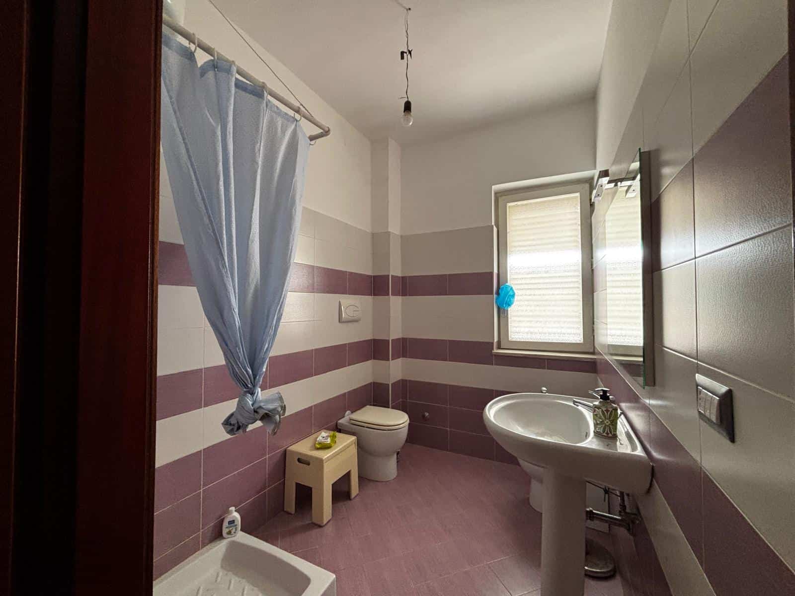 Lavabo e servizi igienici in bagno moderno con pareti a righe rosa e beige, finestra luminosa e dettagli funzionali, ideale per ambienti di comfort e stile.