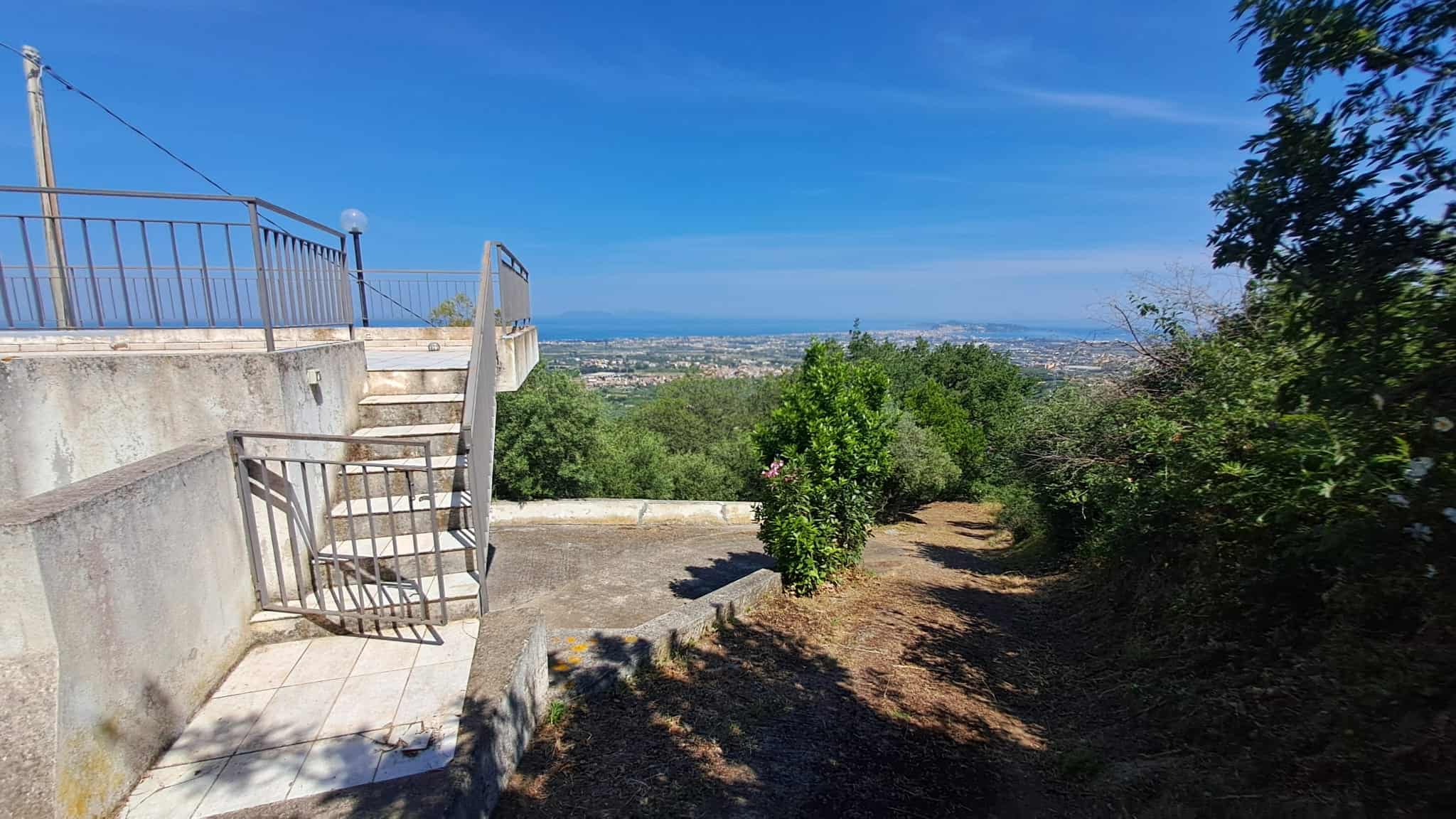 Vista panoramica dal terrazzo con scale in metallo, alberi verdi e paesaggio urbano sullo sfondo, cielo sereno e luminoso, ideale per immobili e proprietà residenziali in vendita o affitto.