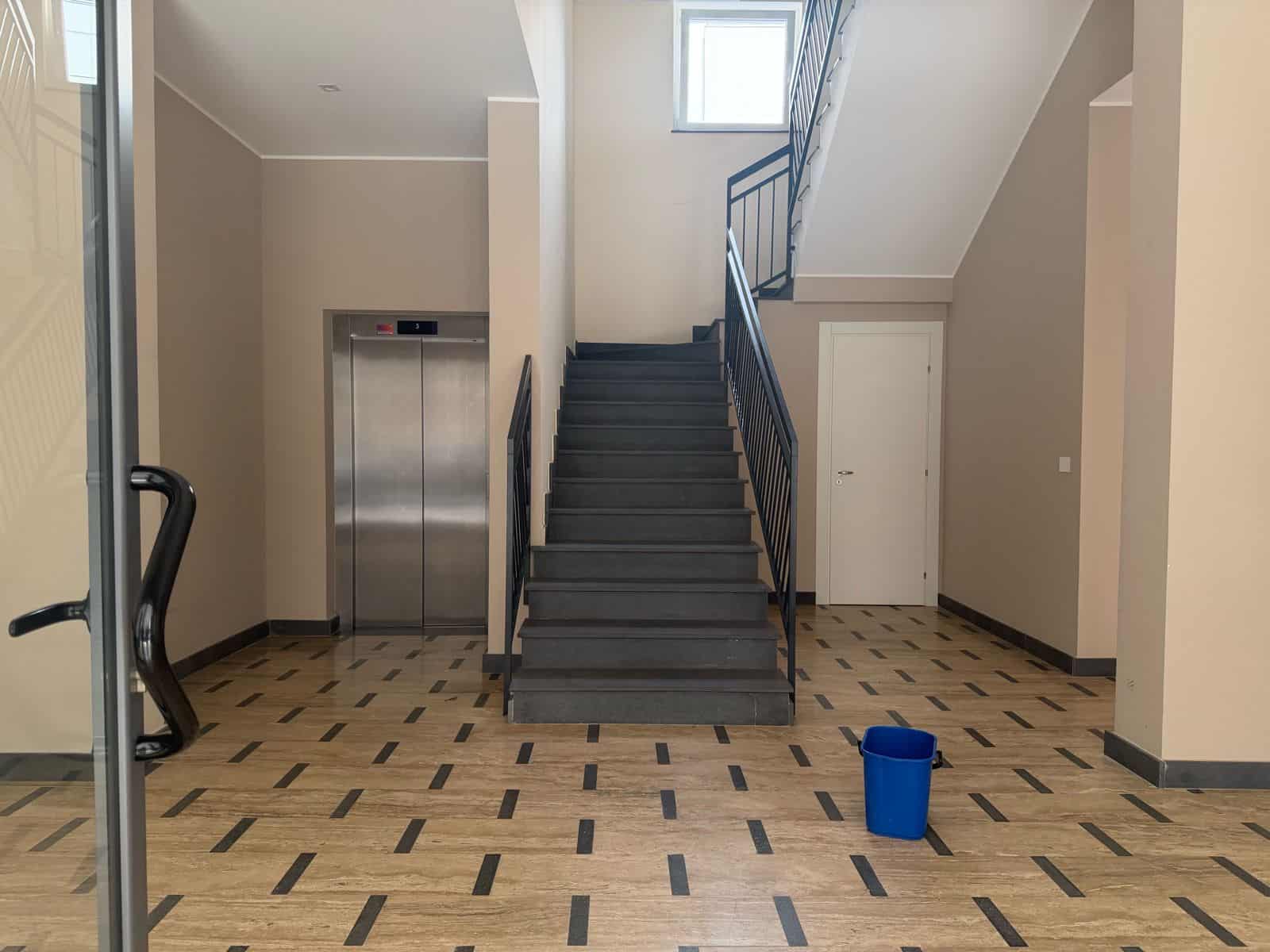 Ascensore e scala in ambiente moderno con pavimento in legno e pareti beige, illuminato da luce naturale da finestra sopra le scale, con secchio blu sul pavimento.