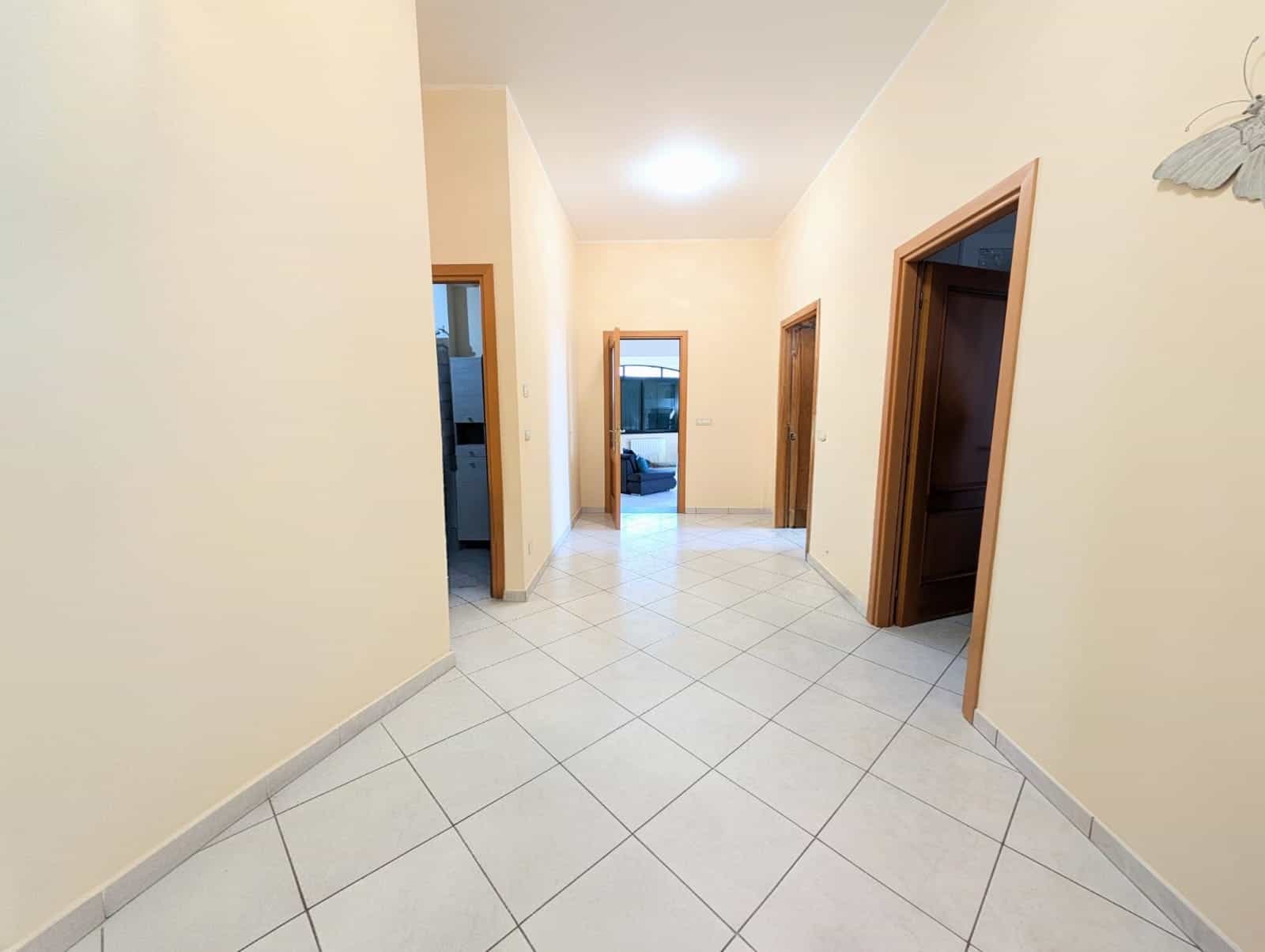 Corridoio interno di una casa con pareti beige, porte in legno e pavimento in piastrelle bianche, illuminato da luce soffusa.