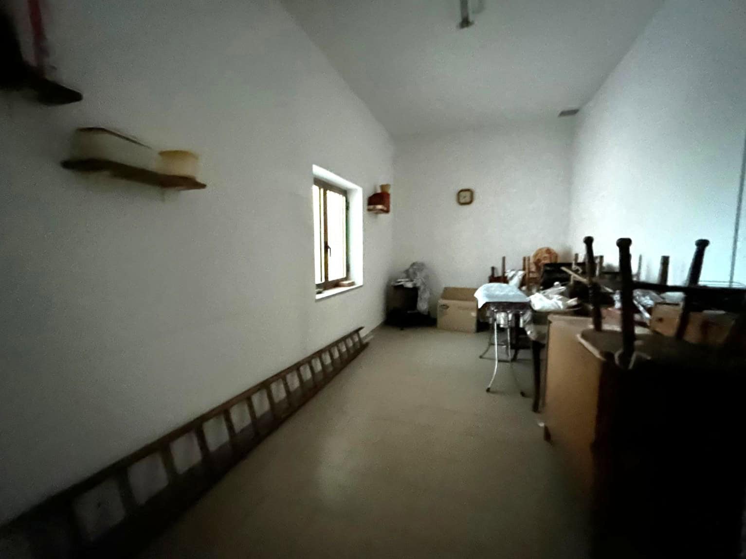 Interno di una stanza vuota con pareti bianche, una finestra e mobili impilati, ambiente semplice e poco arredato.