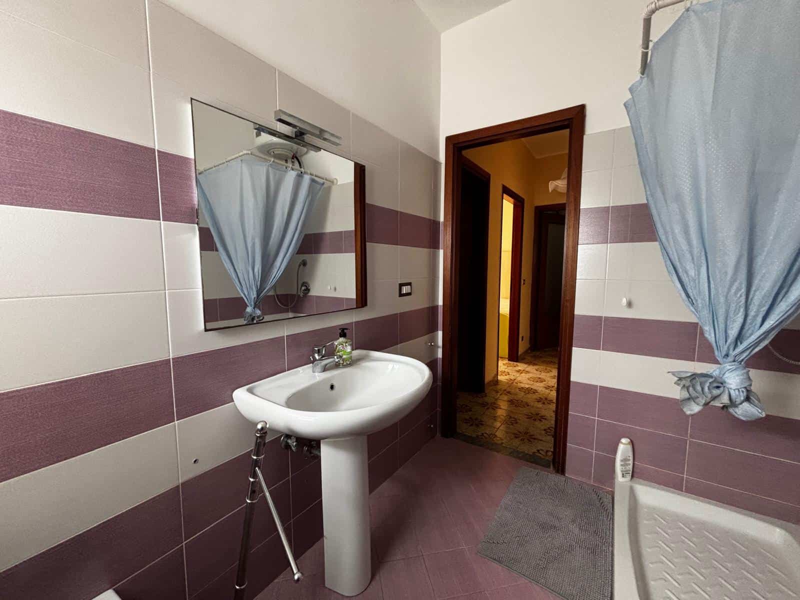 Lavabo e specchio in bagno con pareti a righe viola e beige, porta aperta su corridoio, tende azzurre e pavimento in piastrelle.
