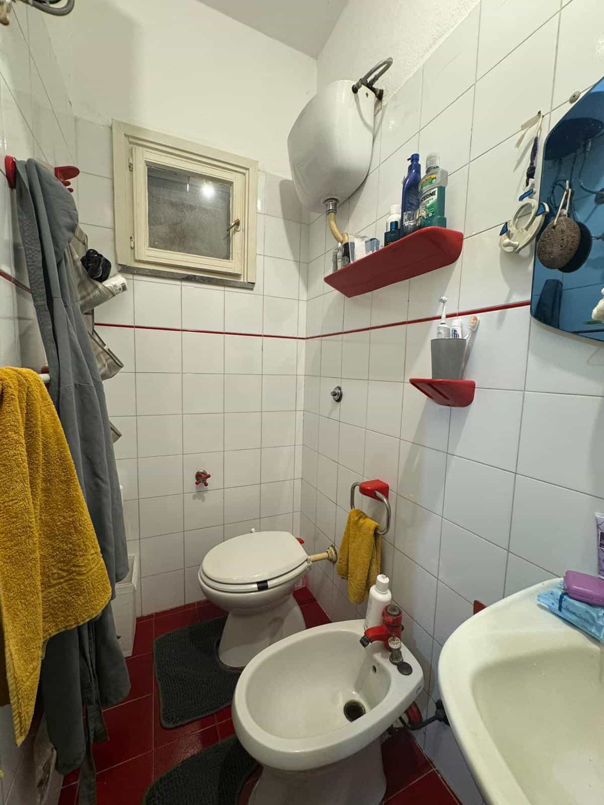 Bagno compatto con WC e bidet, pareti rivestite in piastrelle bianche con dettagli rossi, accessori da bagno e prodotti per l'igiene, spazio funzionale e ben organizzato.