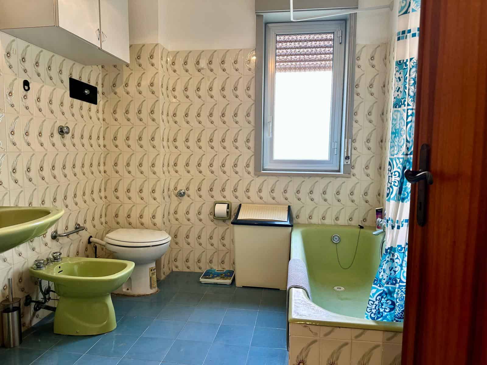 Lavabo e bidet verde in bagno con pareti rivestite di piastrelle beige con motivo a onde, finestra con tende blu e bianche, e vasca da bagno con tendina a motivi blu.