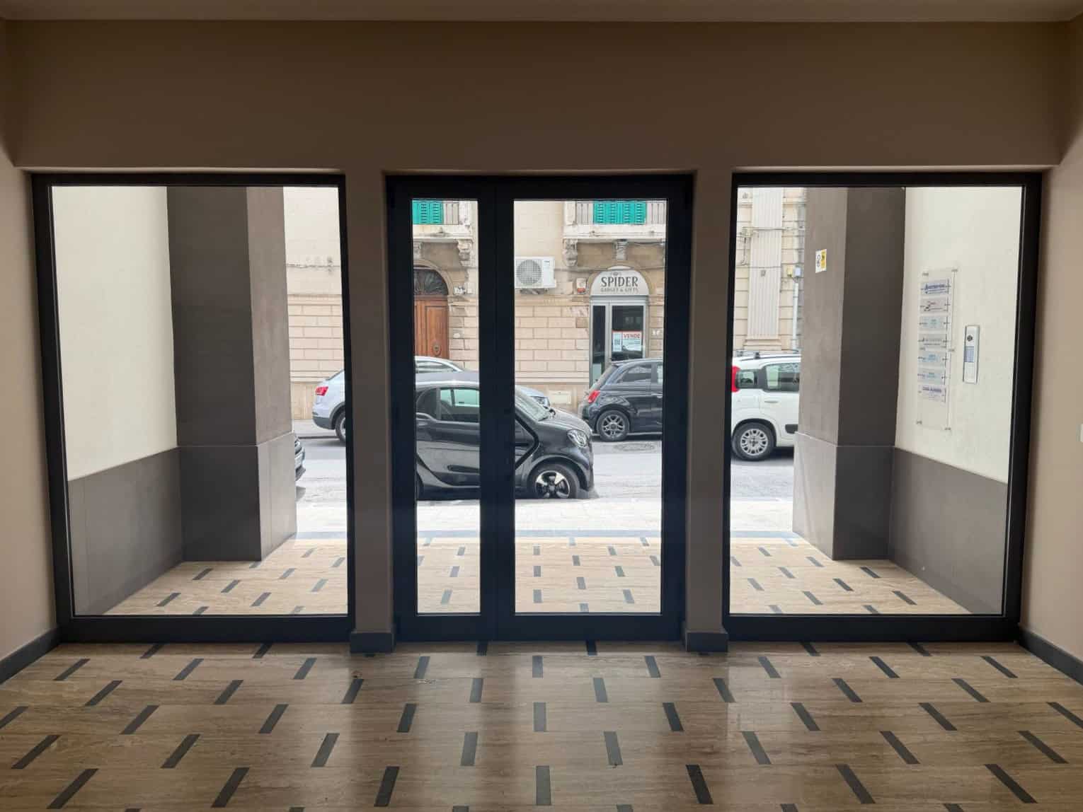 Porta d'ingresso in vetro con cornice nera, vista sulla strada con auto parcheggiate e edifici storici sullo sfondo, ingresso luminoso e moderno.