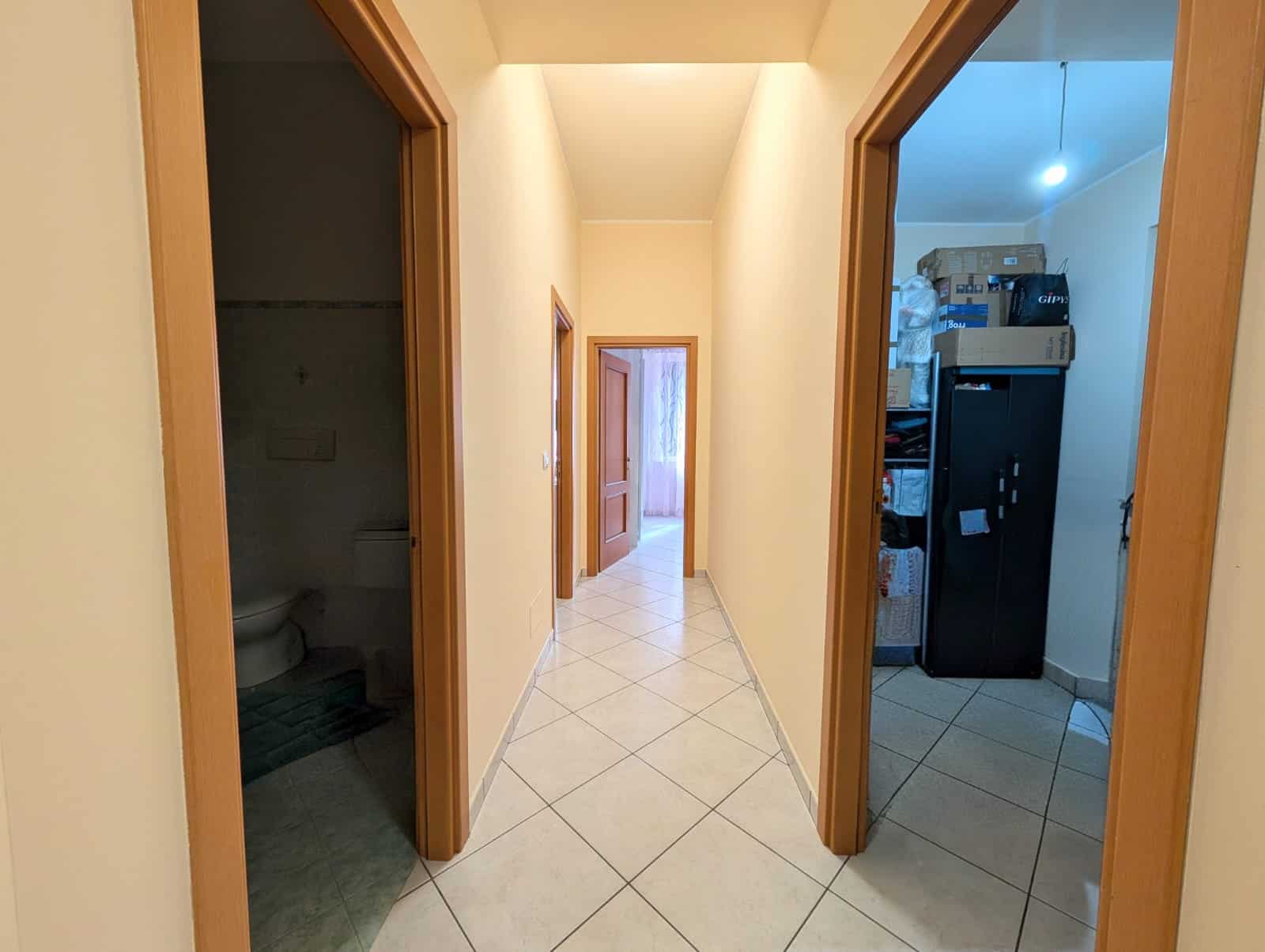 Ampia vista di un corridoio interno di una casa, con pareti color crema e pavimento in piastrelle chiare, che collega diverse stanze, tra cui un bagno e una cucina, con un ambiente luminoso e accoglie.