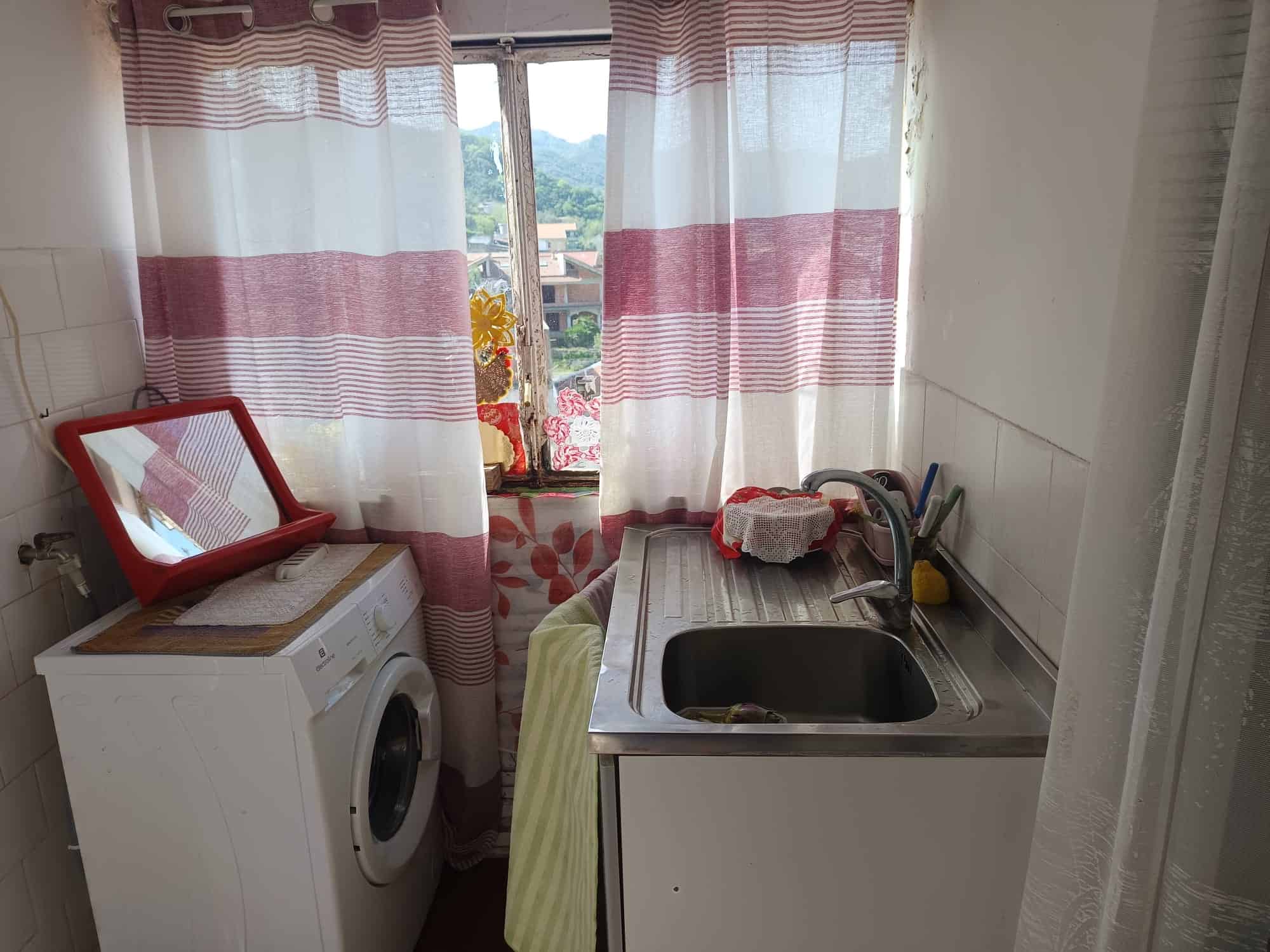 Lavatrice e lavabo in una cucina con tende a righe rosse e bianche, vista su un paesaggio naturale con colline e case.