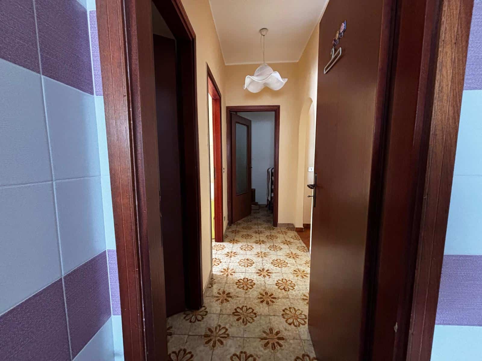 Ampia vista di un corridoio interno di una casa con pavimento in piastrelle floreali, pareti beige e porte in legno scuro, illuminato da una lampada sospesa. Ideale per ambienti residenziali e ristrut.