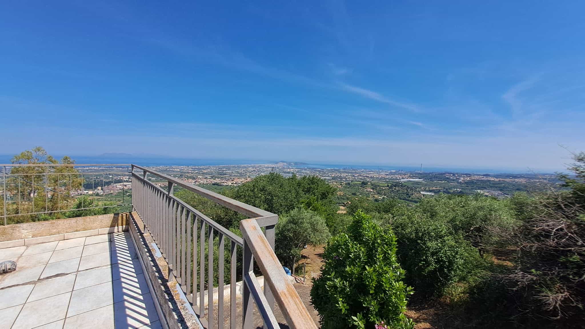 Vista panoramica dal balcone con ringhiera in metallo, che mostra un paesaggio verde e urbano sotto un cielo azzurro, ideale per annunci immobiliari e promozione di immobili con vista.