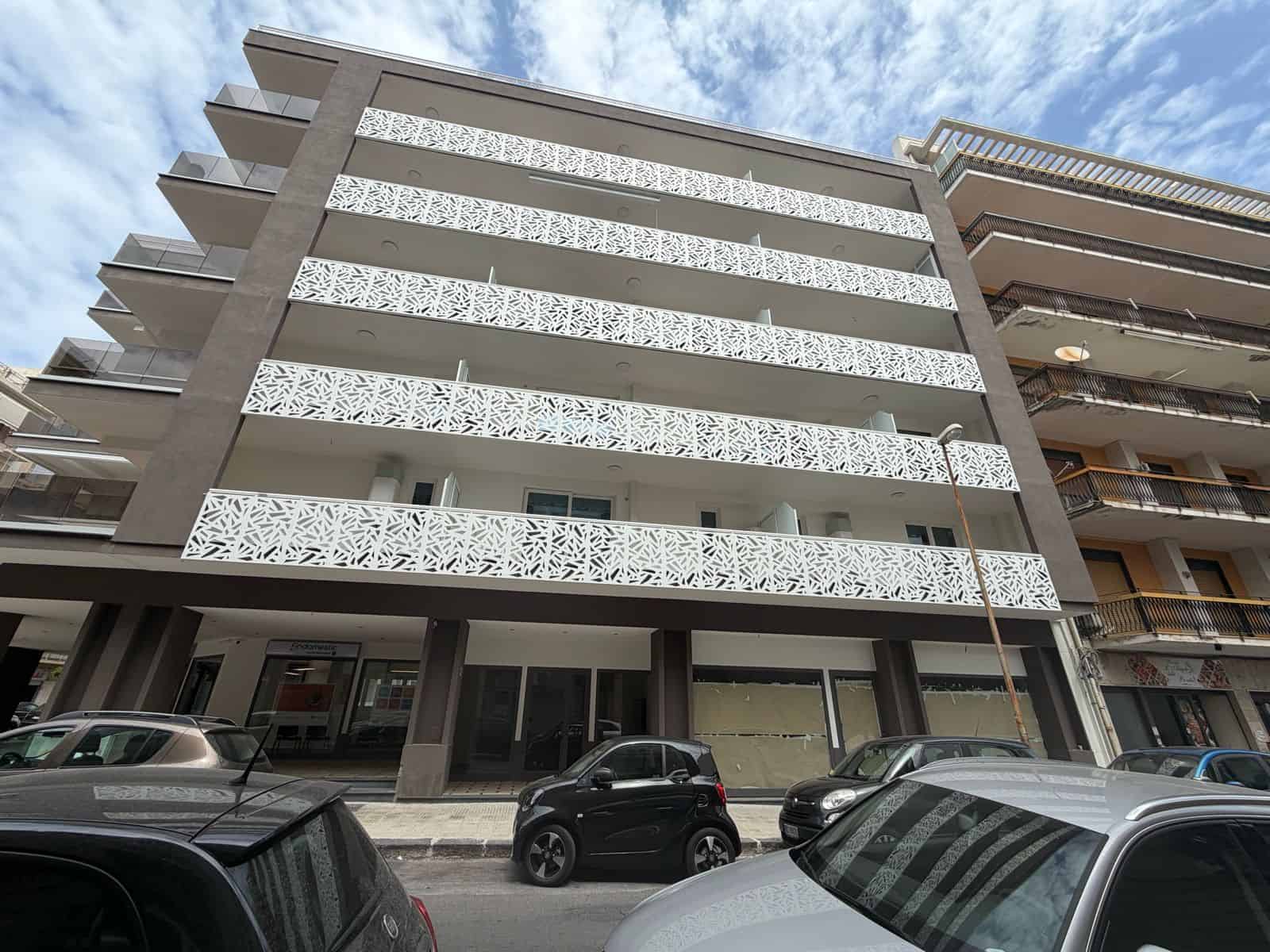 Elegante edificio residenziale moderno con balconi decorativi in ferro battuto, situato in zona urbana con parcheggi frontali, sotto un cielo sereno.