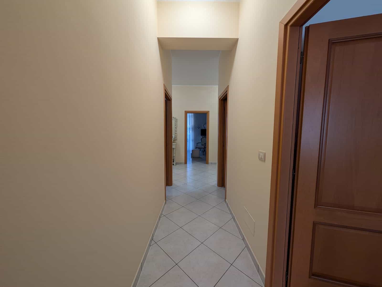 Ampia corridoio interno di casa con pareti beige e porte in legno scuro, illuminato da luce naturale, con pavimento in piastrelle chiare, ideale per ambienti residenziali moderni.