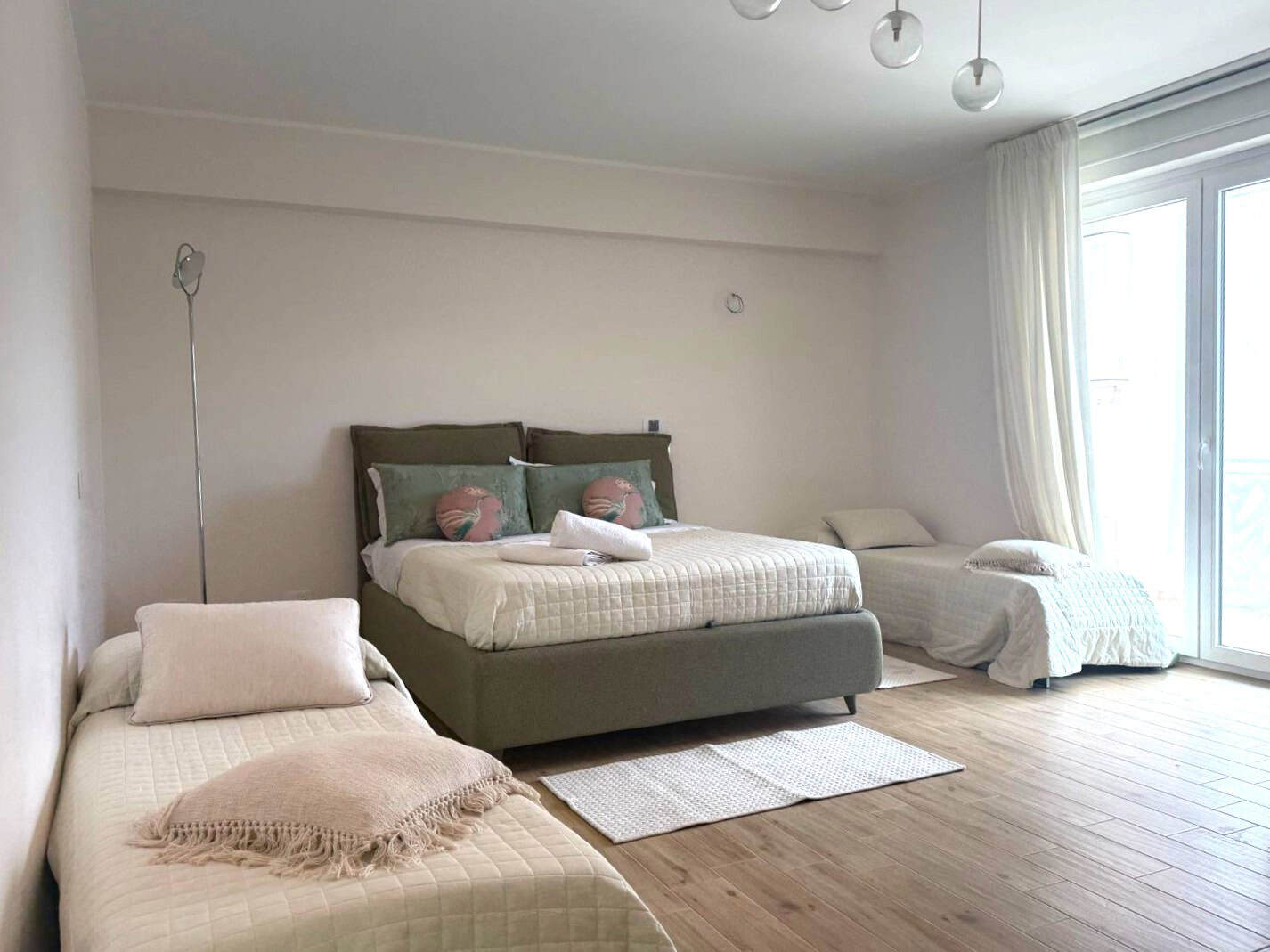 Letto matrimoniale con due cuscini decorativi in una camera moderna e luminosa, con pareti bianche e pavimento in legno chiaro, vicino a grandi finestre con tende bianche.