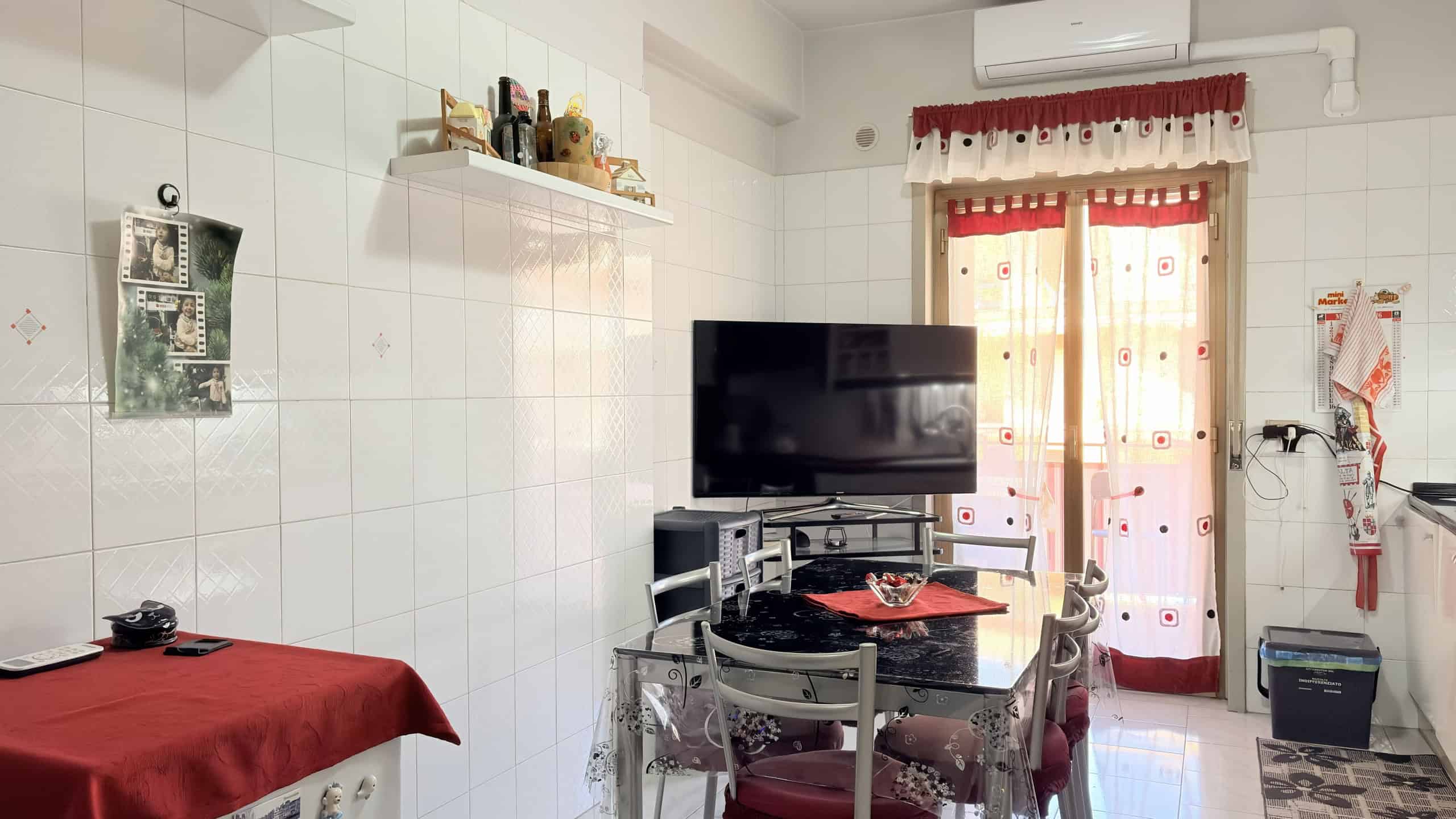Ampio soggiorno con pareti bianche, televisore a schermo piatto, tavolo rotondo con sedie, finestra con tende rosse e bianche, decorazioni e oggetti domestici, ambiente luminoso e accogliente.