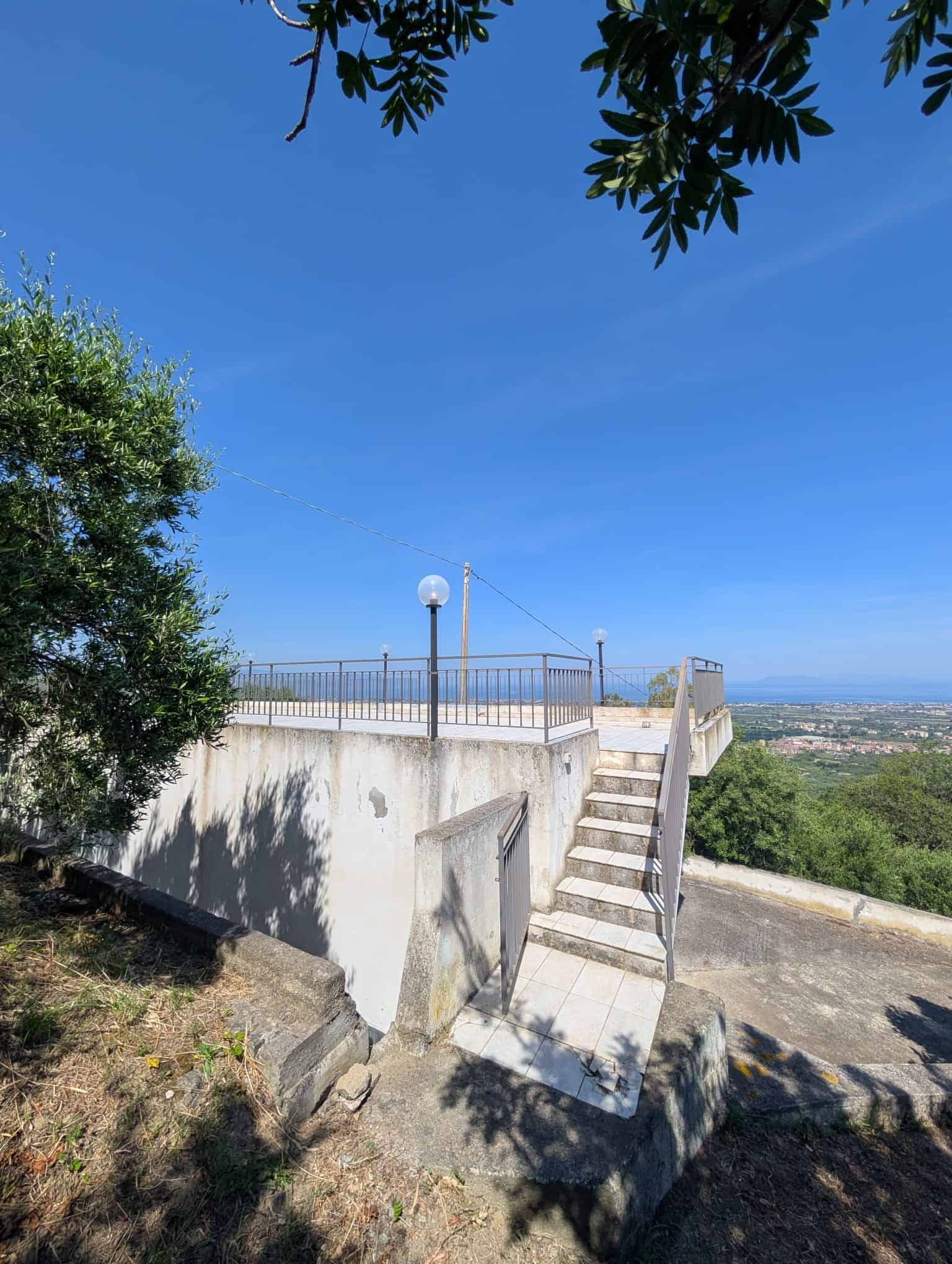 Viale panoramico con vista sulla città e sul paesaggio circostante, terrazza con ringhiera in metallo, lampione e scale in cemento, ambiente esterno soleggiato e tranquillo, ideale per relax e momenti.