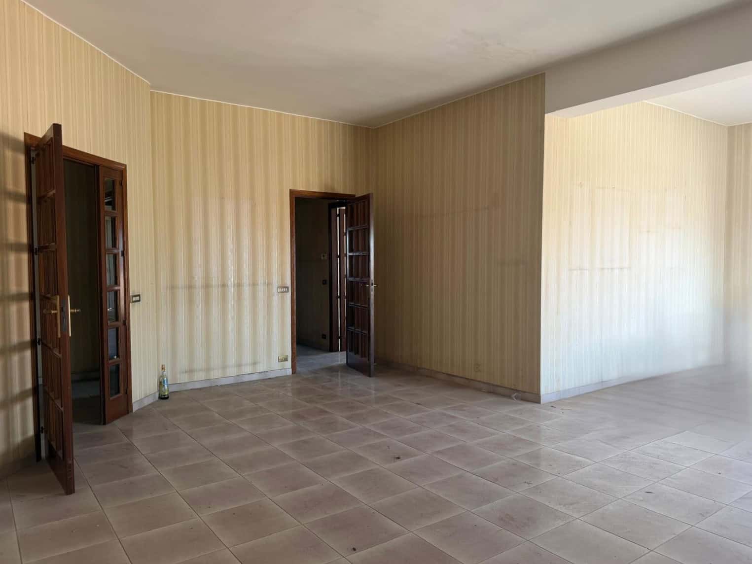 Ampia stanza vuota con pareti beige e pavimento in piastrelle, ideale per ristrutturazione o arredamento personalizzato, perfetta per creare un ambiente accogliente e funzionale.