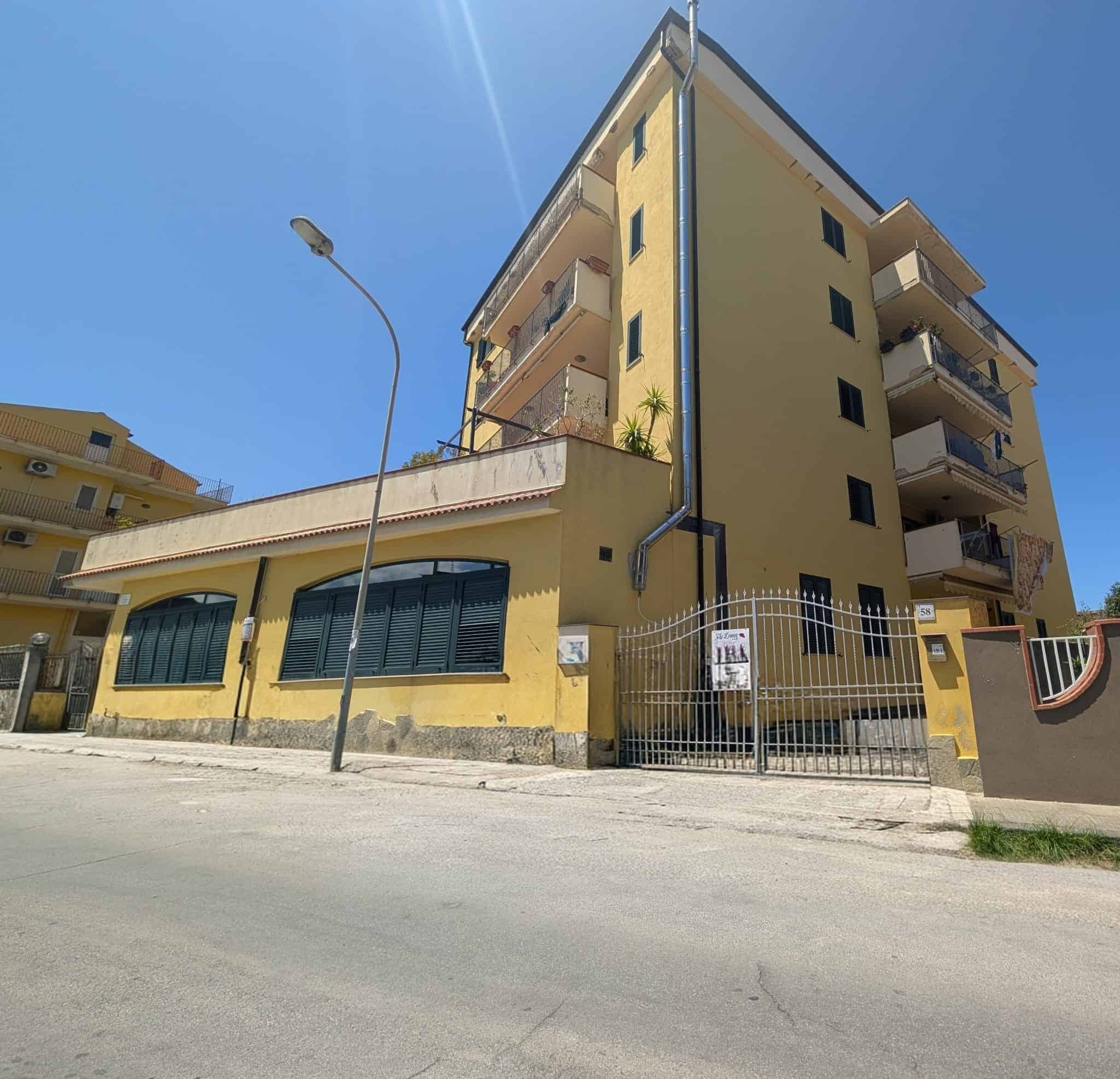 Ampio edificio residenziale giallo con balconi e finestre, situato in zona urbana soleggiata con strada e lampione in primo piano, vista chiara e luminosa.