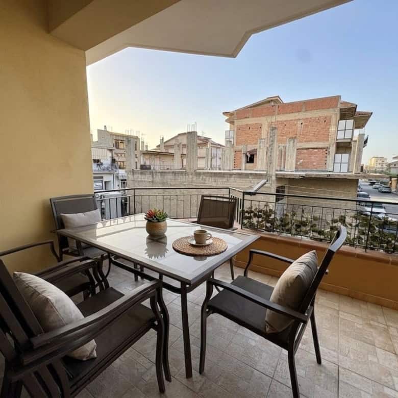 Balcone di un appartamento con tavolo e sedie, vista su edifici residenziali in un quartiere urbano, luce naturale, atmosfera accogliente e moderna.