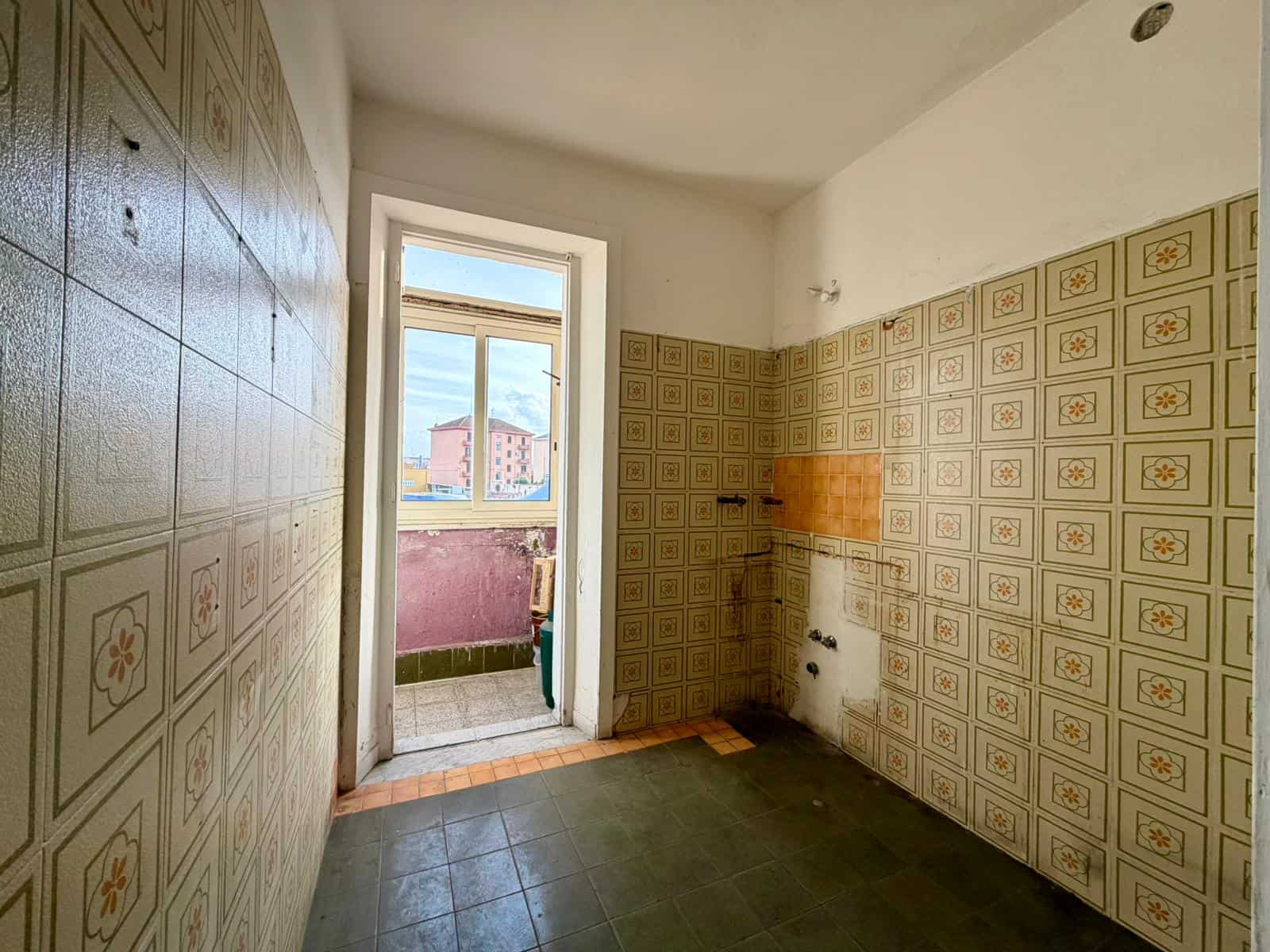 Cucina vuota con pareti rivestite di piastrelle decorative, porta finestra che conduce a un balcone con vista sulla città, pavimento in piastrelle scure.
