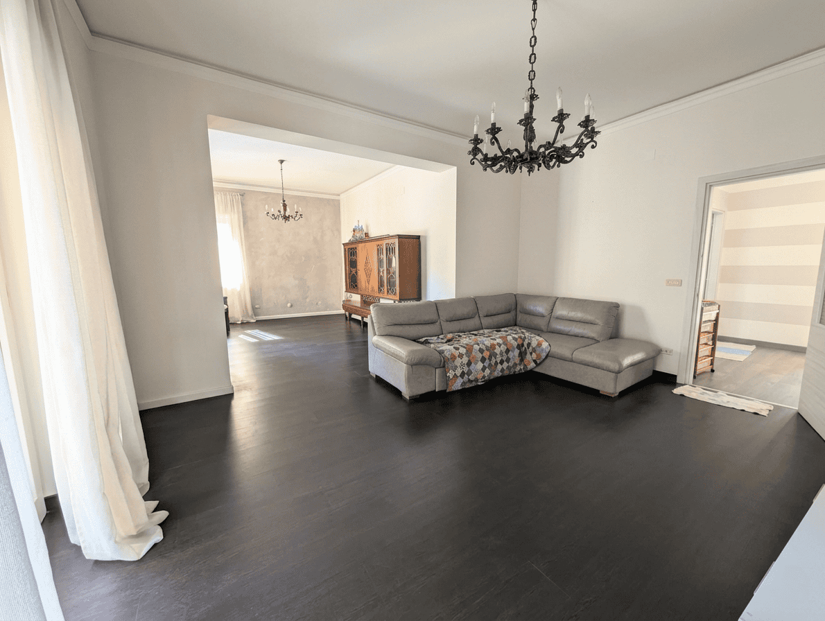 Interno di un soggiorno spazioso con pavimento in legno scuro, divano angolare grigio, lampadario antico e pareti bianche, ambiente luminoso e accogliente.