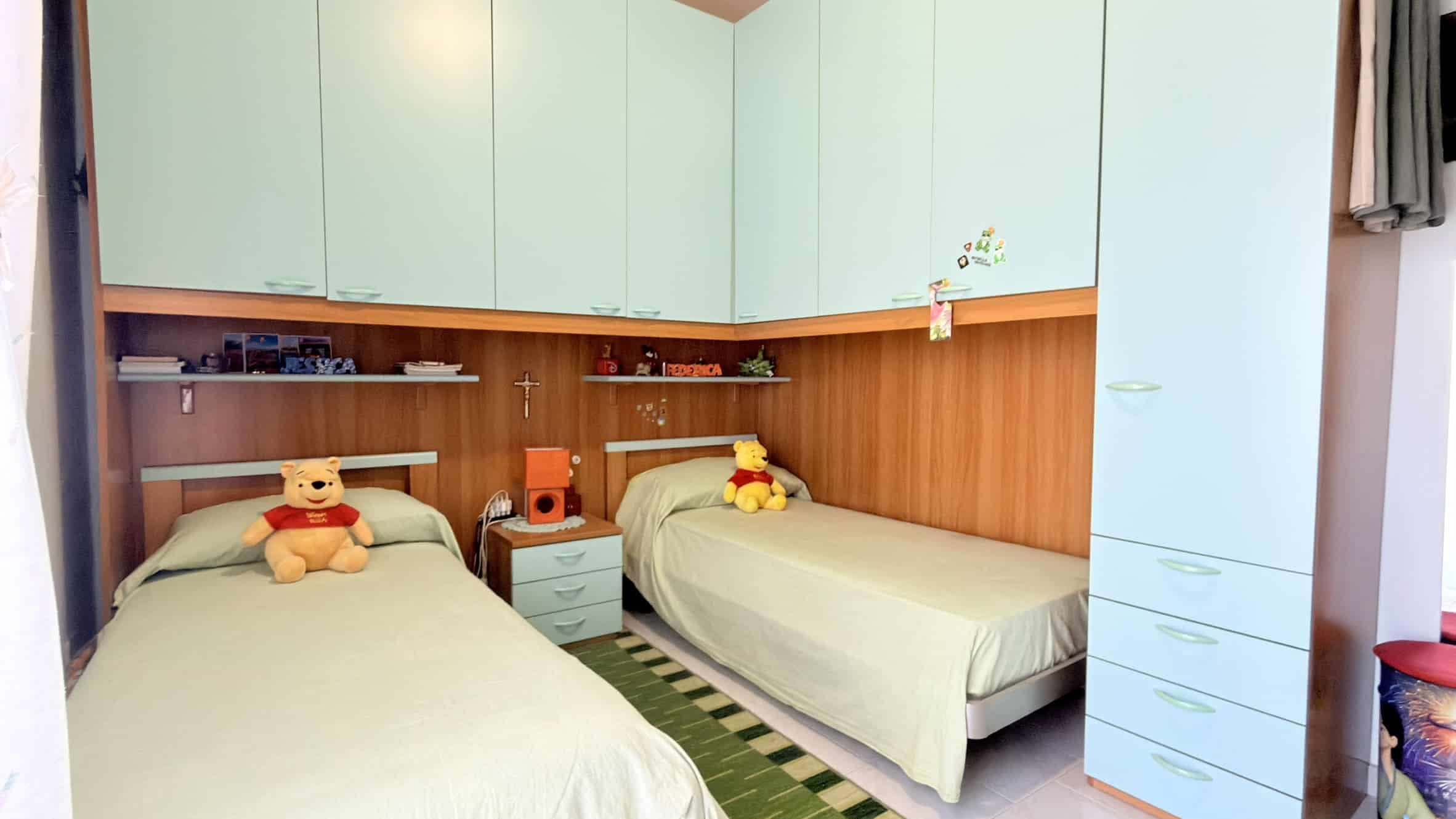 Letto singolo con testiera in legno e armadio a muro in una camera da letto moderna, decorata con teddy bear e accessori colorati, ideale per ambienti giovani e funzionali.