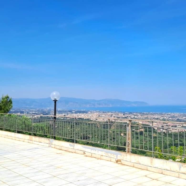 Vista panoramica sulla città e sul mare dal terrazzo di una casa con pavimentazione in piastrelle e lampioni decorativi, immersa nel verde e con un ampio orizzonte.