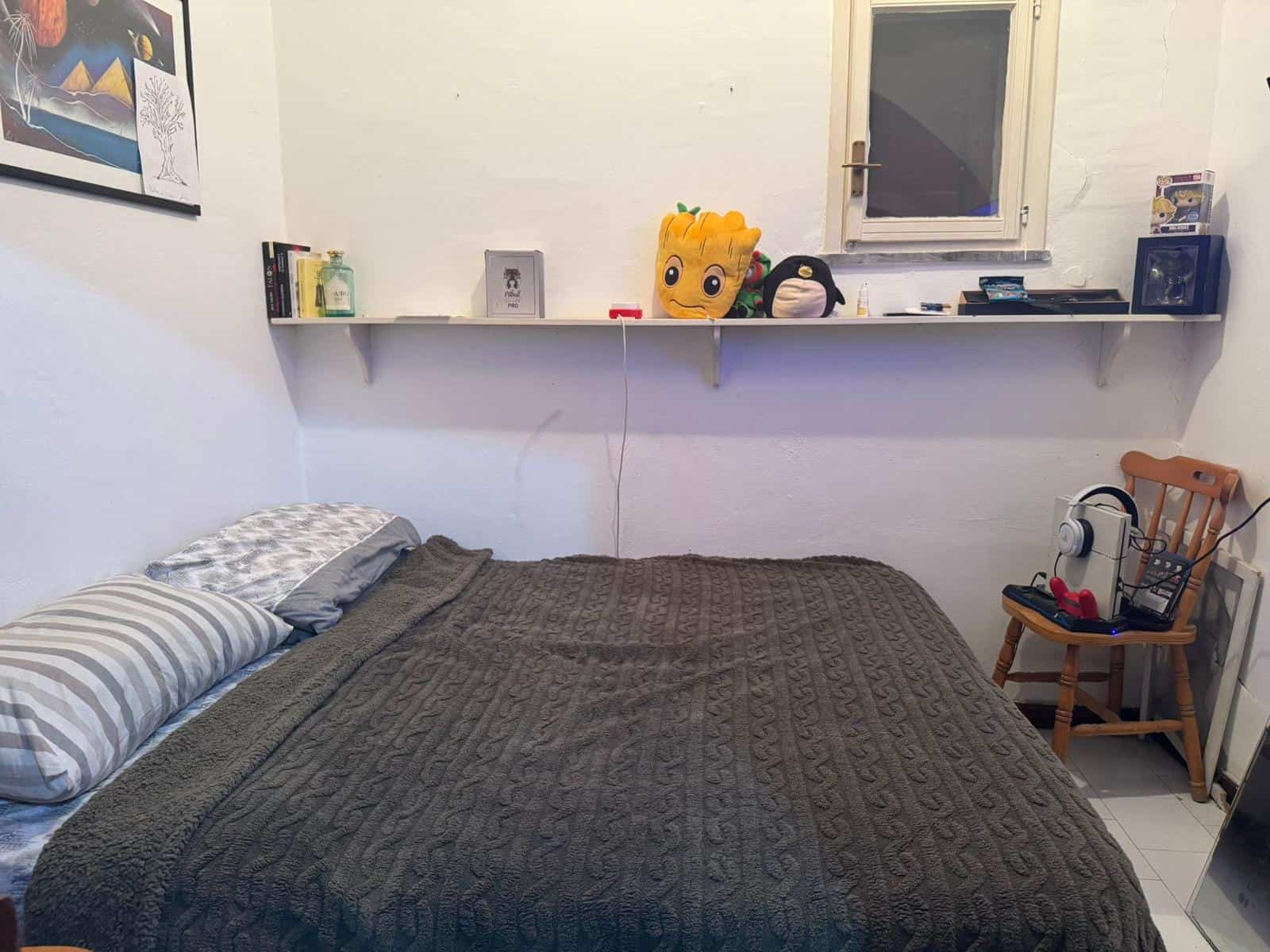 Camera da letto semplice con letto matrimoniale, mensola con decorazioni e oggetti vari, sedia in legno, ambiente domestico accogliente e funzionale.