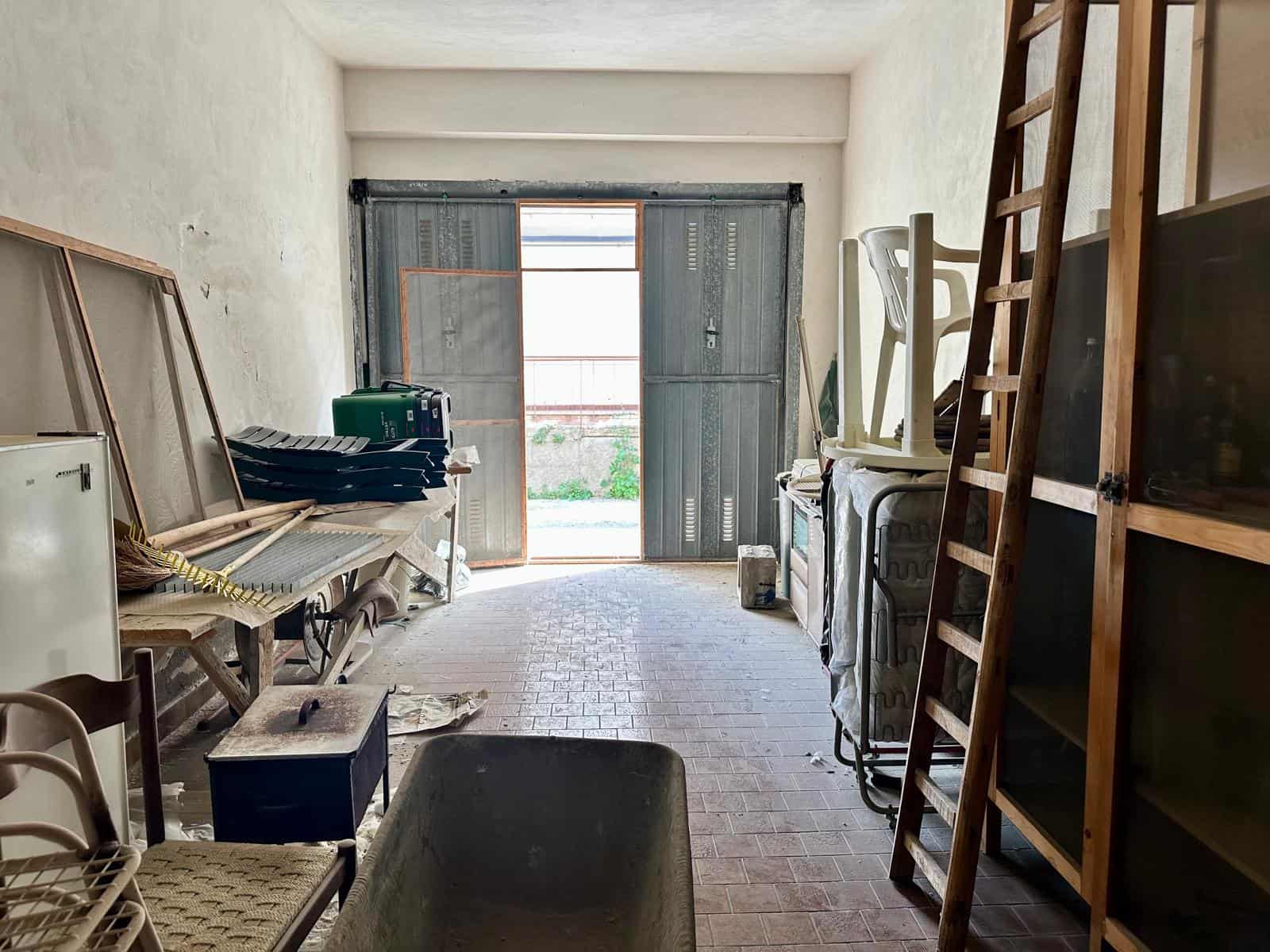 Interno di un magazzino o deposito con scaffali, attrezzi e materiali di stoccaggio, luce naturale dall'ingresso, ambiente organizzato e funzionale.