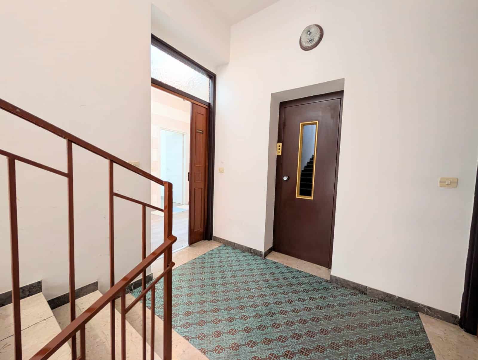 Porta d'ingresso e corridoio in un edificio residenziale, con pavimento in piastrelle e pareti bianche, illuminato dalla luce naturale proveniente dalla porta aperta.