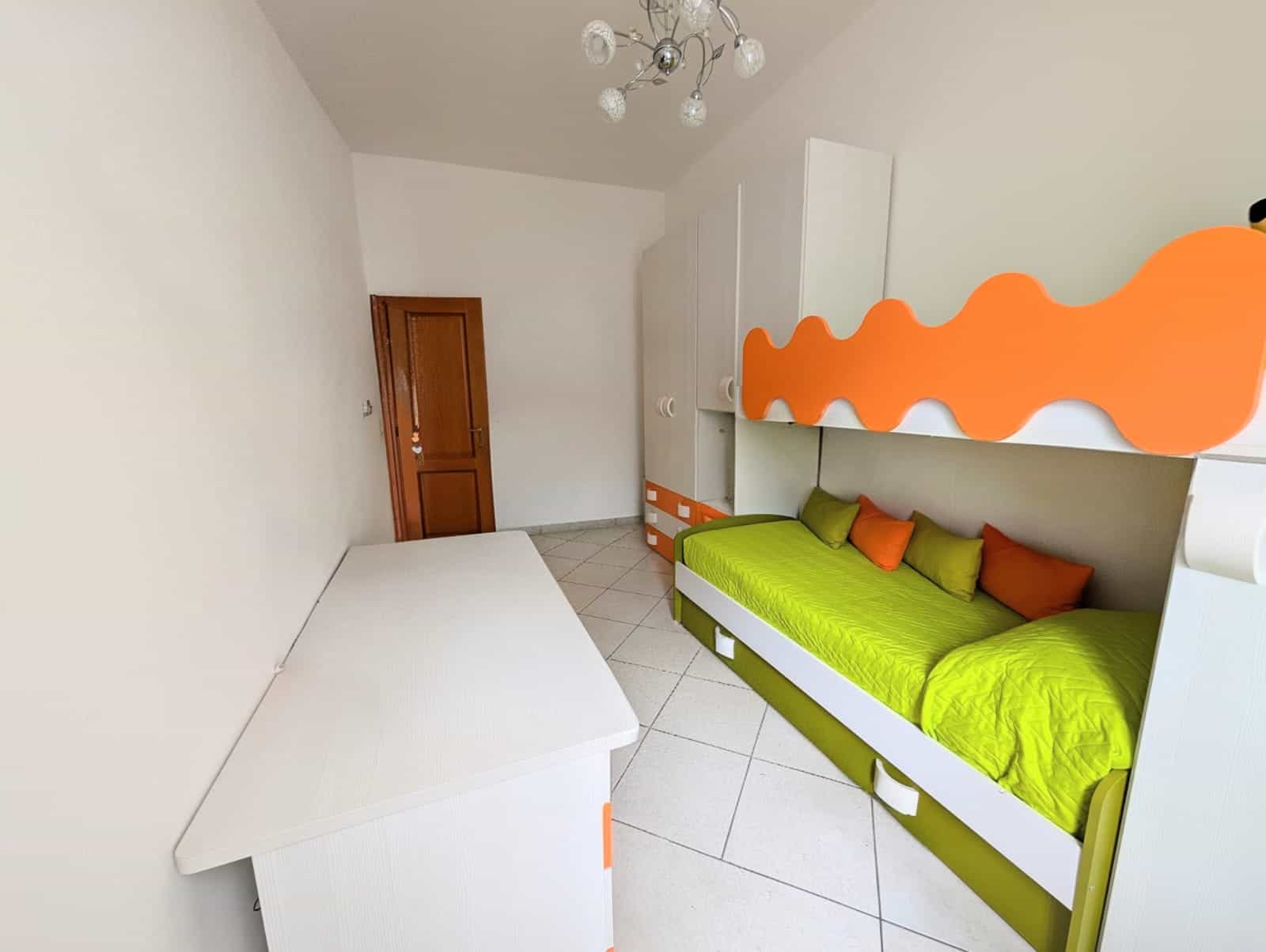Letto singolo con testiera colorata in camera moderna, decorata con elementi arancioni e verdi, illuminata da lampadario a soffitto, ideale per ambienti giovani e vivaci.