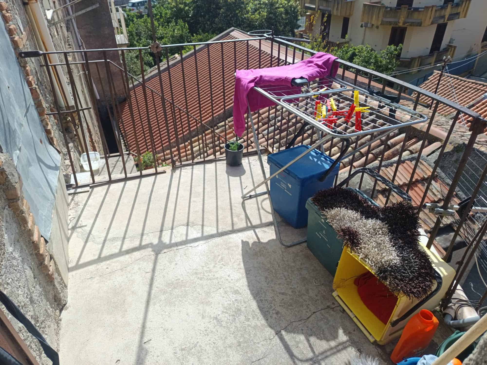 Lavatrice e stendibiancheria sul balcone di un appartamento, con vista sui tetti delle case vicine e sul verde degli alberi circostanti.