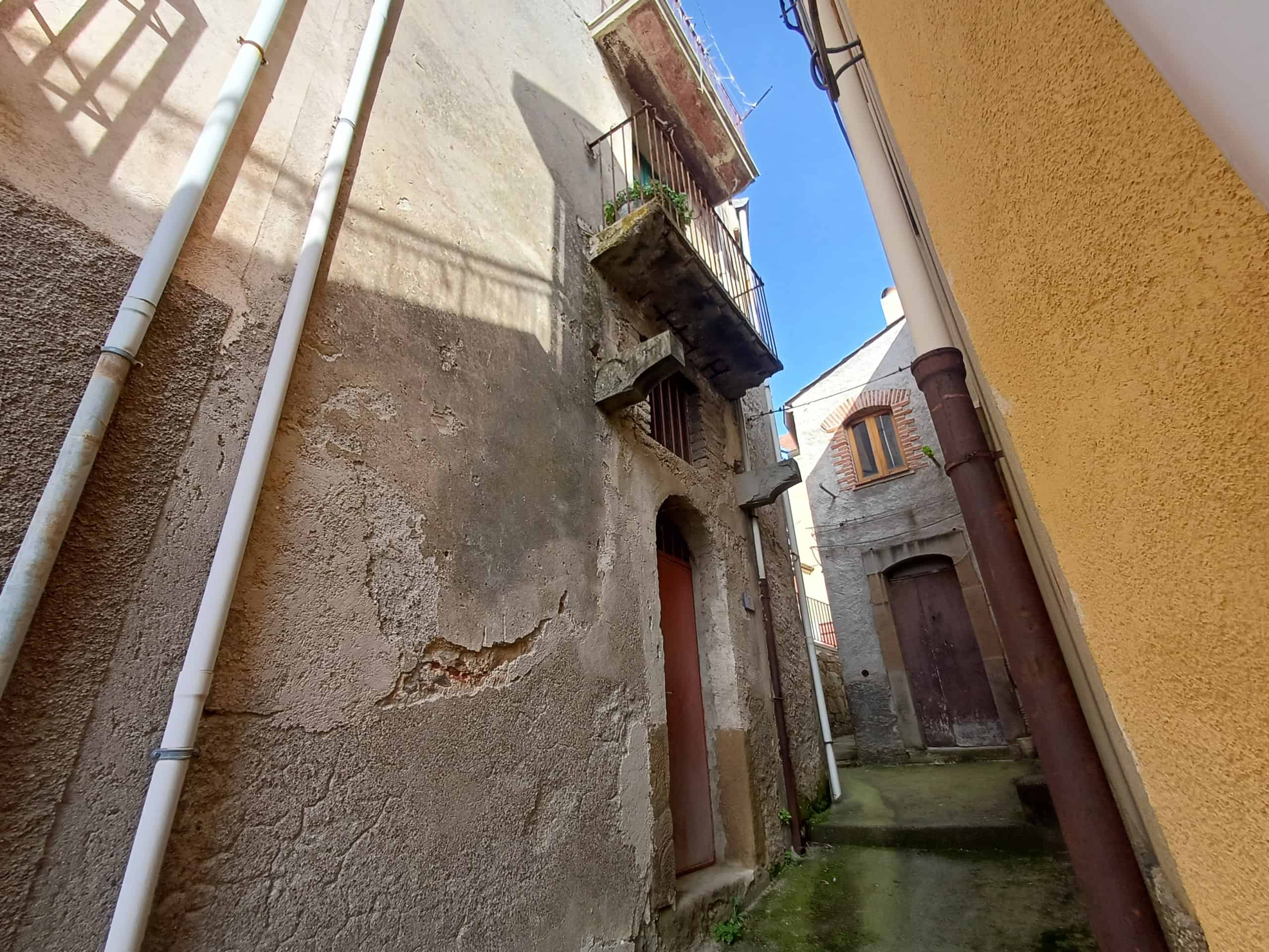 Casa antica in pietra con balcone e piante, situata in un vicolo storico di un borgo italiano.