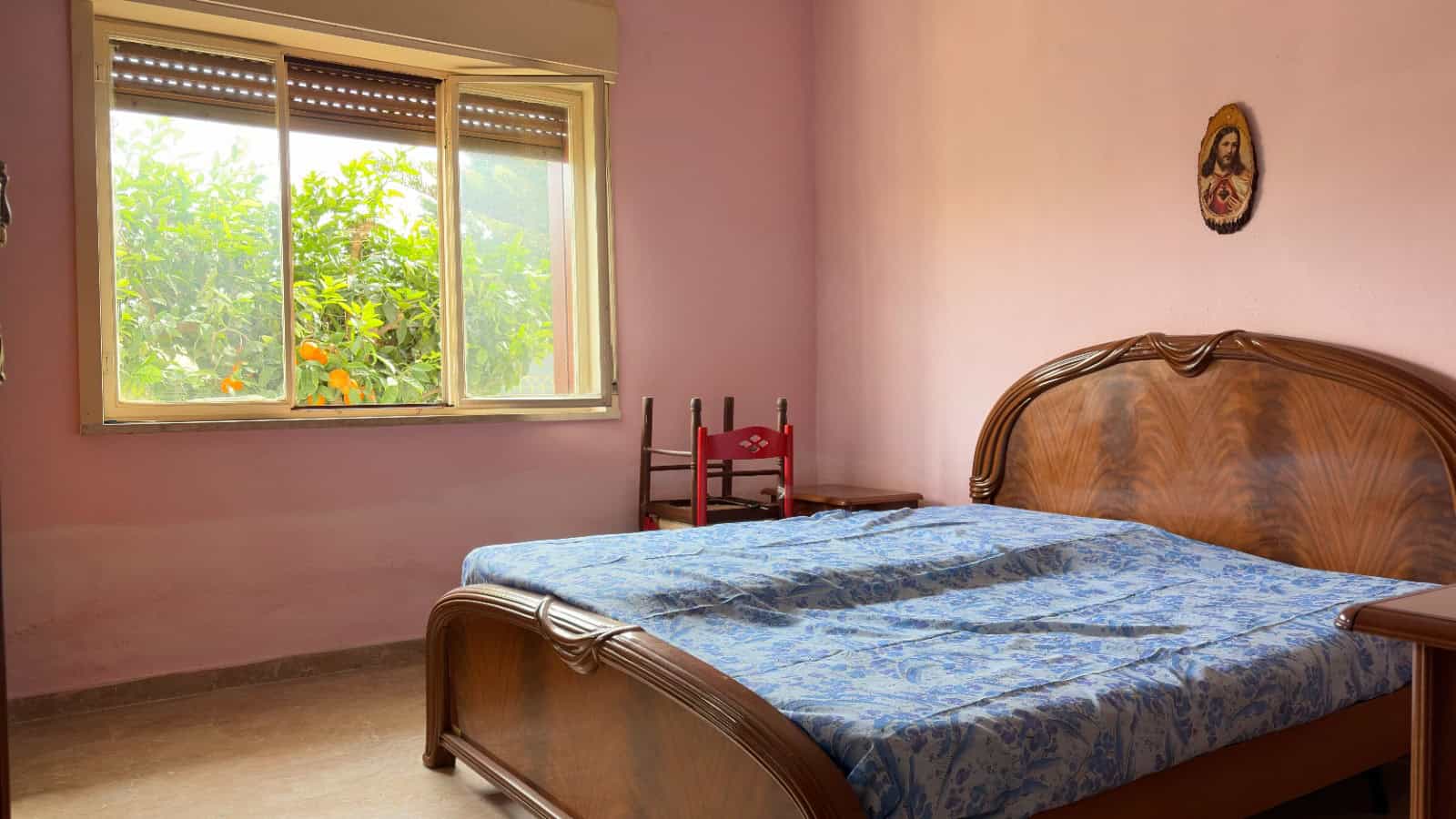 Soggiorno con letto matrimoniale in una stanza luminosa e accogliente, con pareti rosa e finestra che mostra un giardino verde. Ideale per affitti di case o appartamenti su NextCasa.