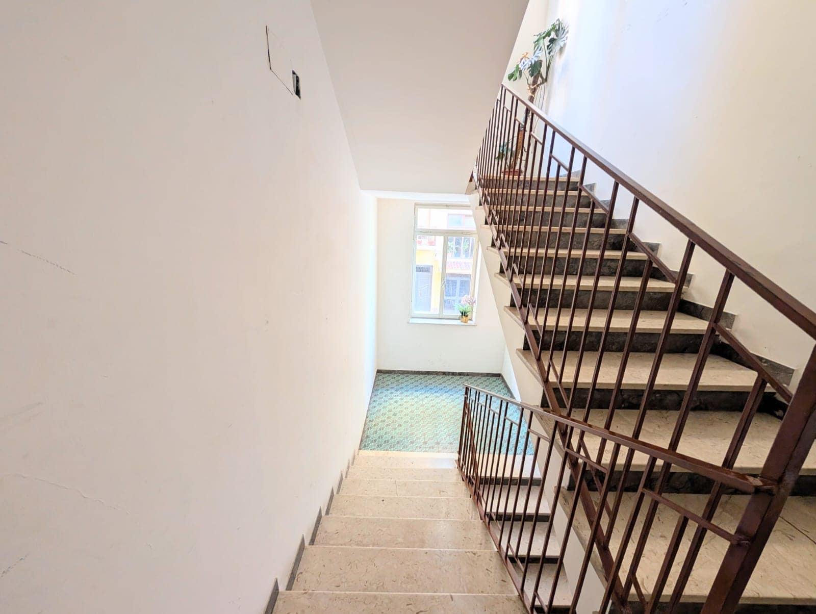 Scalinata interna di un edificio residenziale con ringhiera in metallo e pareti bianche, illuminata dalla luce naturale proveniente da una finestra.