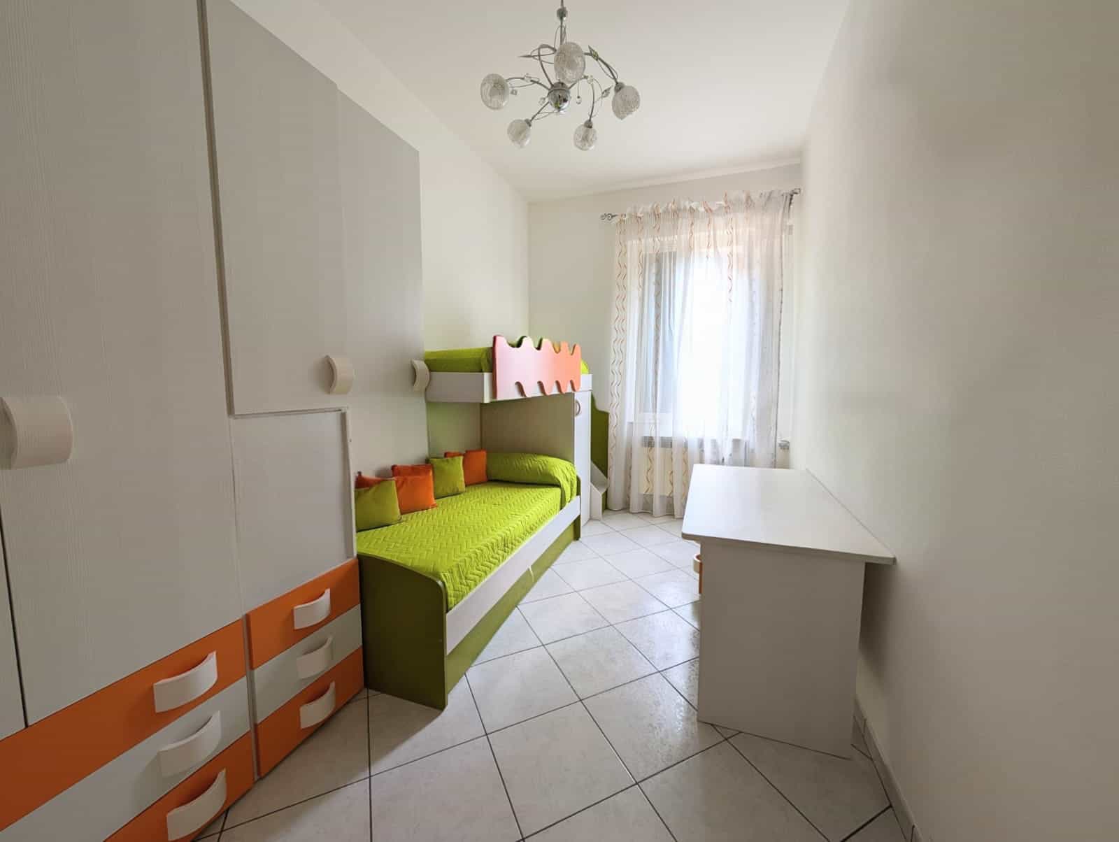 L'immagine mostra una cameretta per bambini con mobili colorati, tra cui un letto a castello verde con dettagli rosa e arancioni, una scrivania bianca e una finestra con tende leggere, in un ambiente.