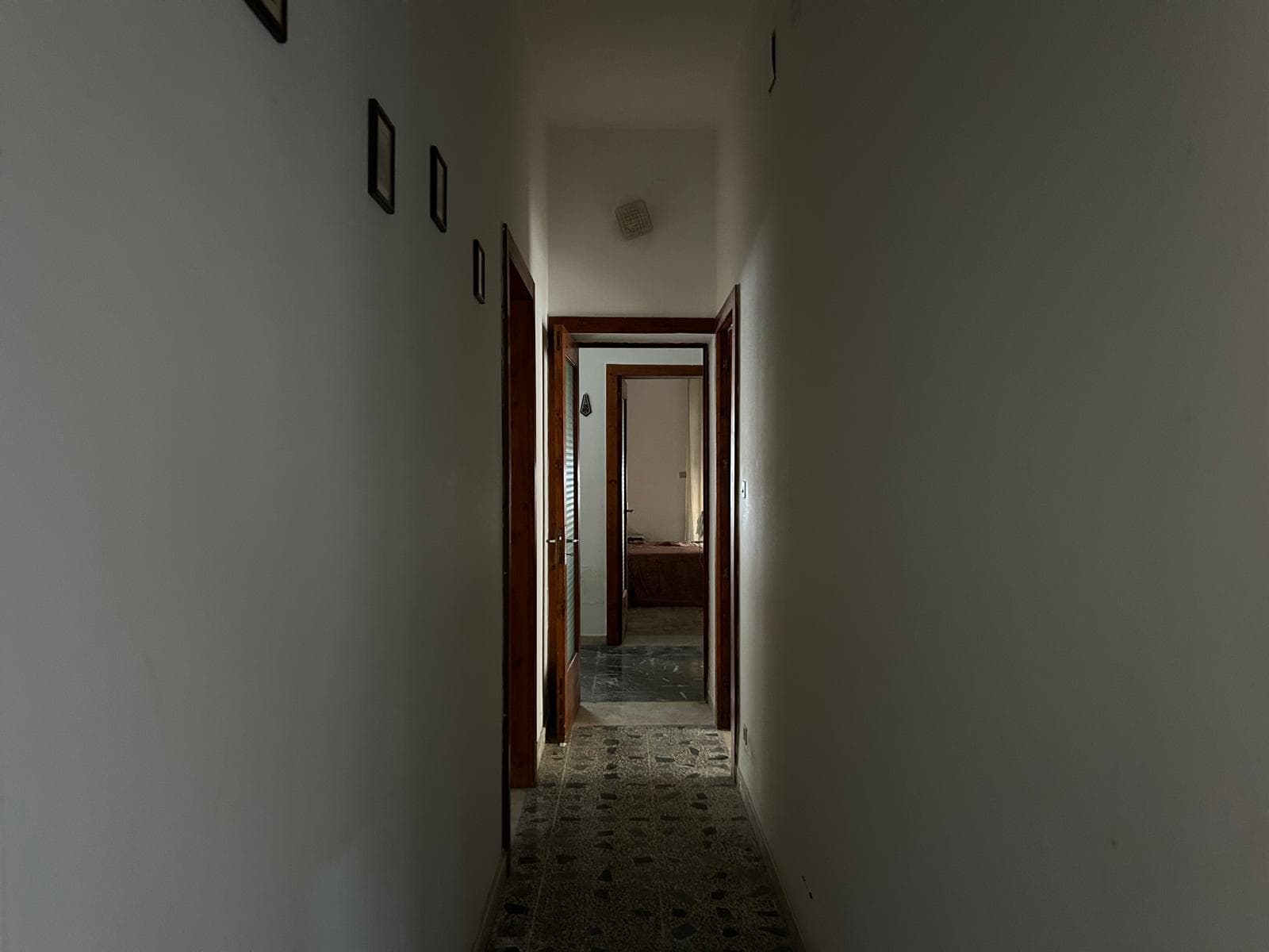 Corridoio interno di un appartamento con pareti bianche, porte in legno e pavimento in piastrelle, vista verso una stanza con arredi.
