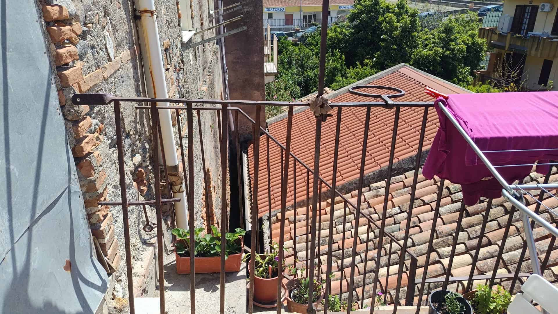 Scorcio di un balcone con ringhiera in ferro battuto, piante in vaso e vista su tetti di tegole rosse e aree verdi circostanti, in un contesto urbano italiano.