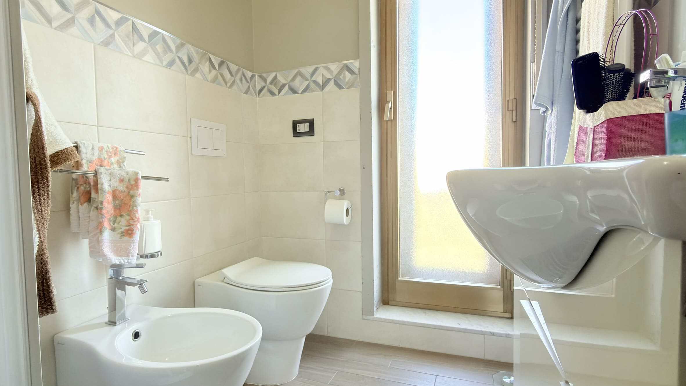 Spazio bagno moderno con lavabo, WC e finestra luminosa, arredato con accessori e asciugamani colorati, ideale per ambienti di comfort e funzionalità in case e appartamenti.