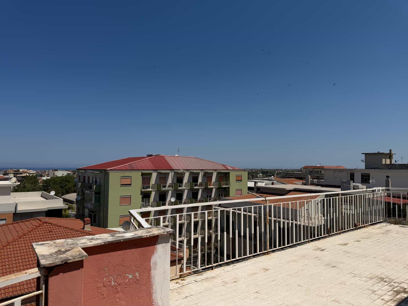 VISTA PANORAMICA di un terrazzo con ringhiera in metallo, affacciato su un quartiere residenziale con edifici di diverse altezze e colori, sotto un cielo sereno e limpido.