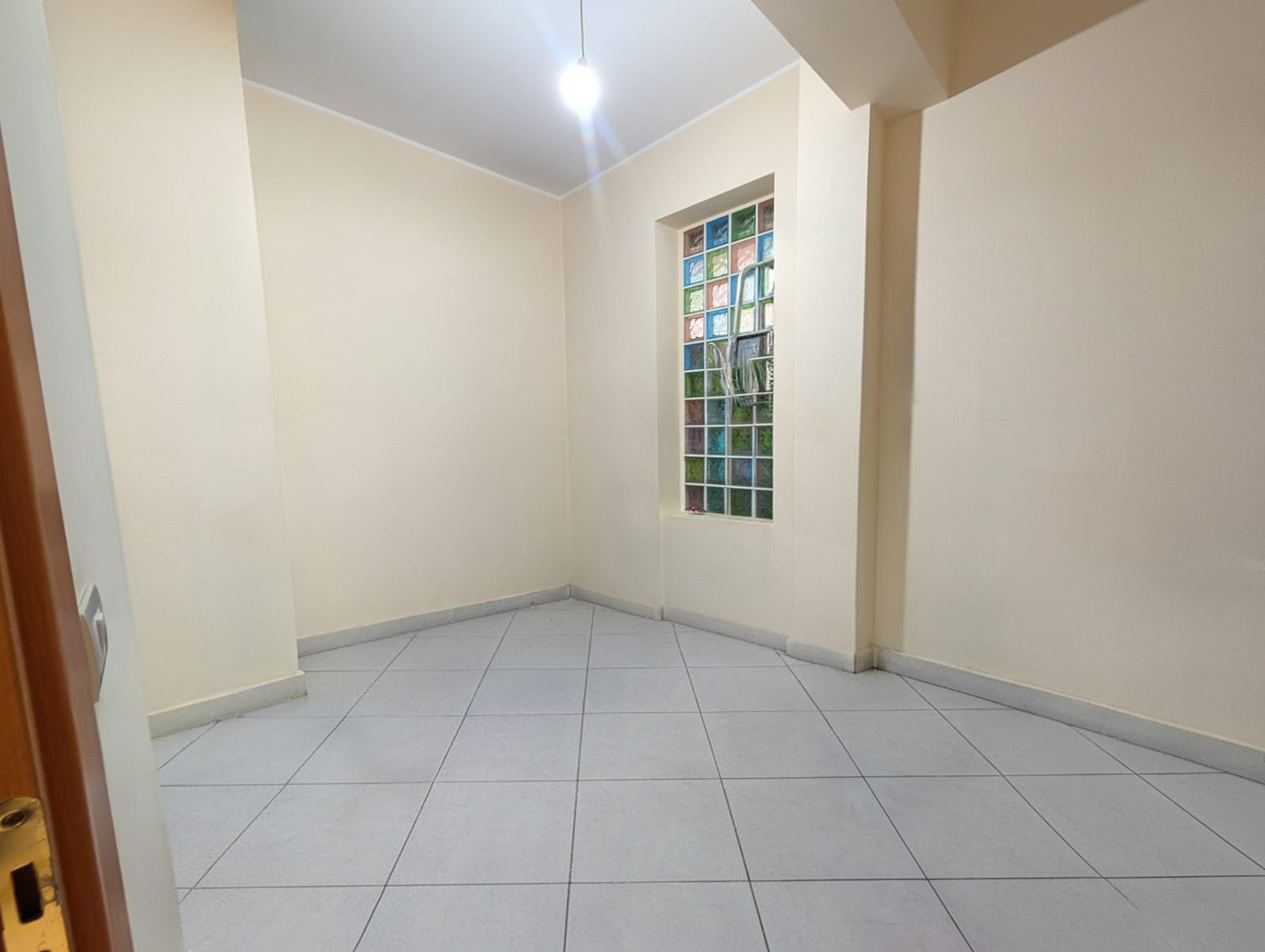 Ampia stanza vuota con pareti beige, finestra con vetri colorati e pavimento in piastrelle grigie, ideale per ristrutturazione o arredamento personalizzato.