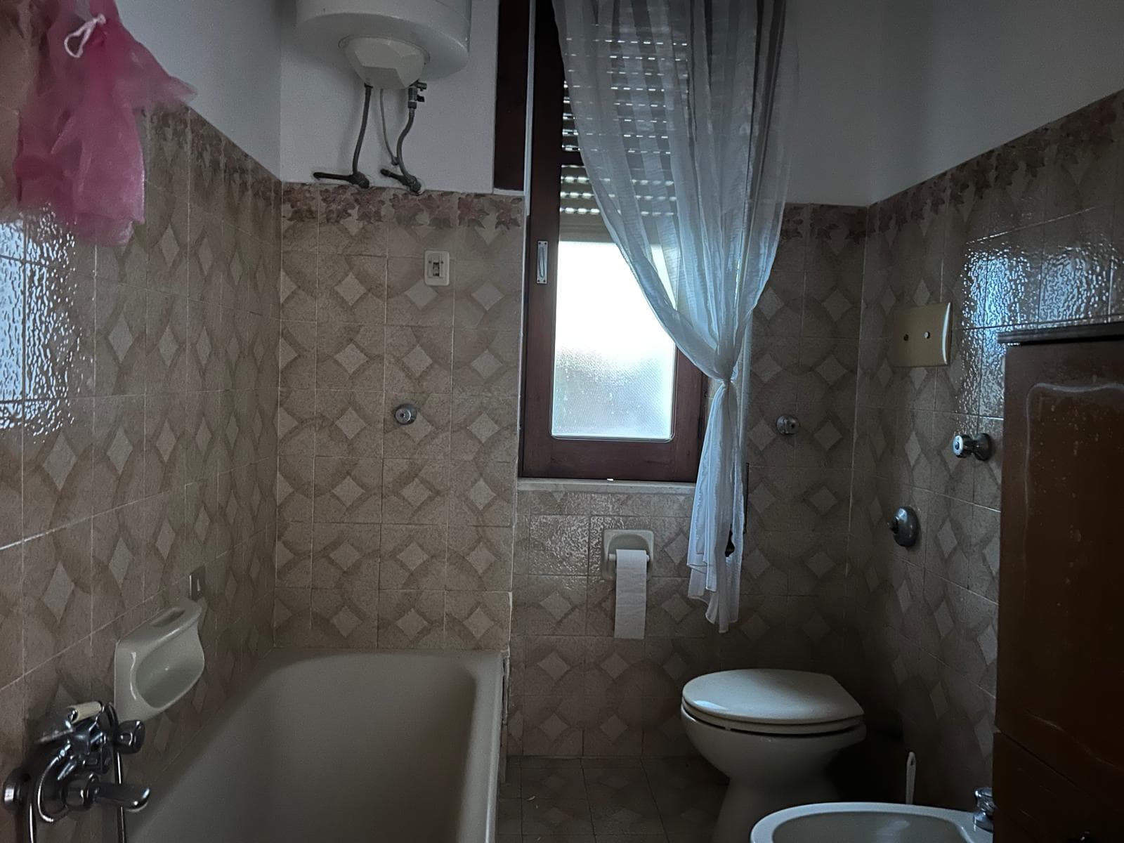 Lavabo e vasca da bagno in un bagno di piccole dimensioni con pareti rivestite in piastrelle beige e finestra con tende leggere.