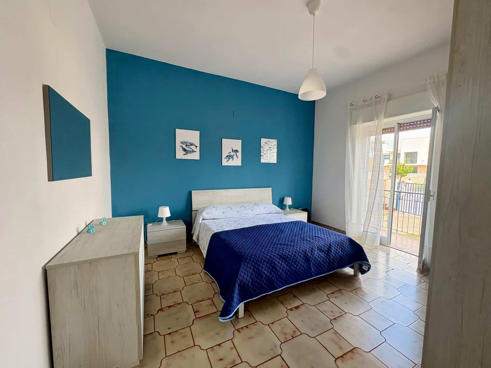 Il letto matrimoniale in una camera moderna con parete blu, decorazioni marine e accessori minimalisti, con accesso a un balcone soleggiato, ideale per soggiorni rilassanti e confortevoli.