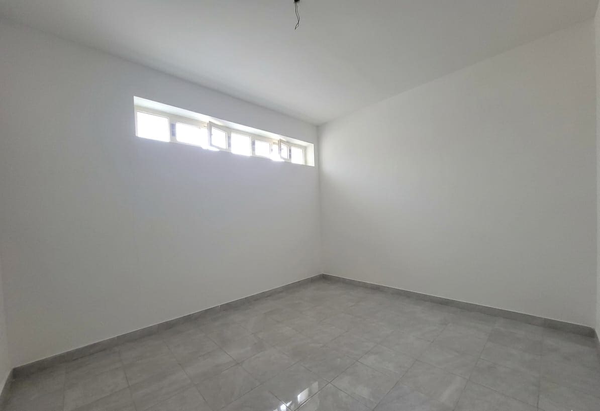 Ampia stanza vuota con pareti bianche, finestra alta e naturale illuminazione, pavimento in piastrelle grigie, ideale per arredamento moderno e personalizzato.