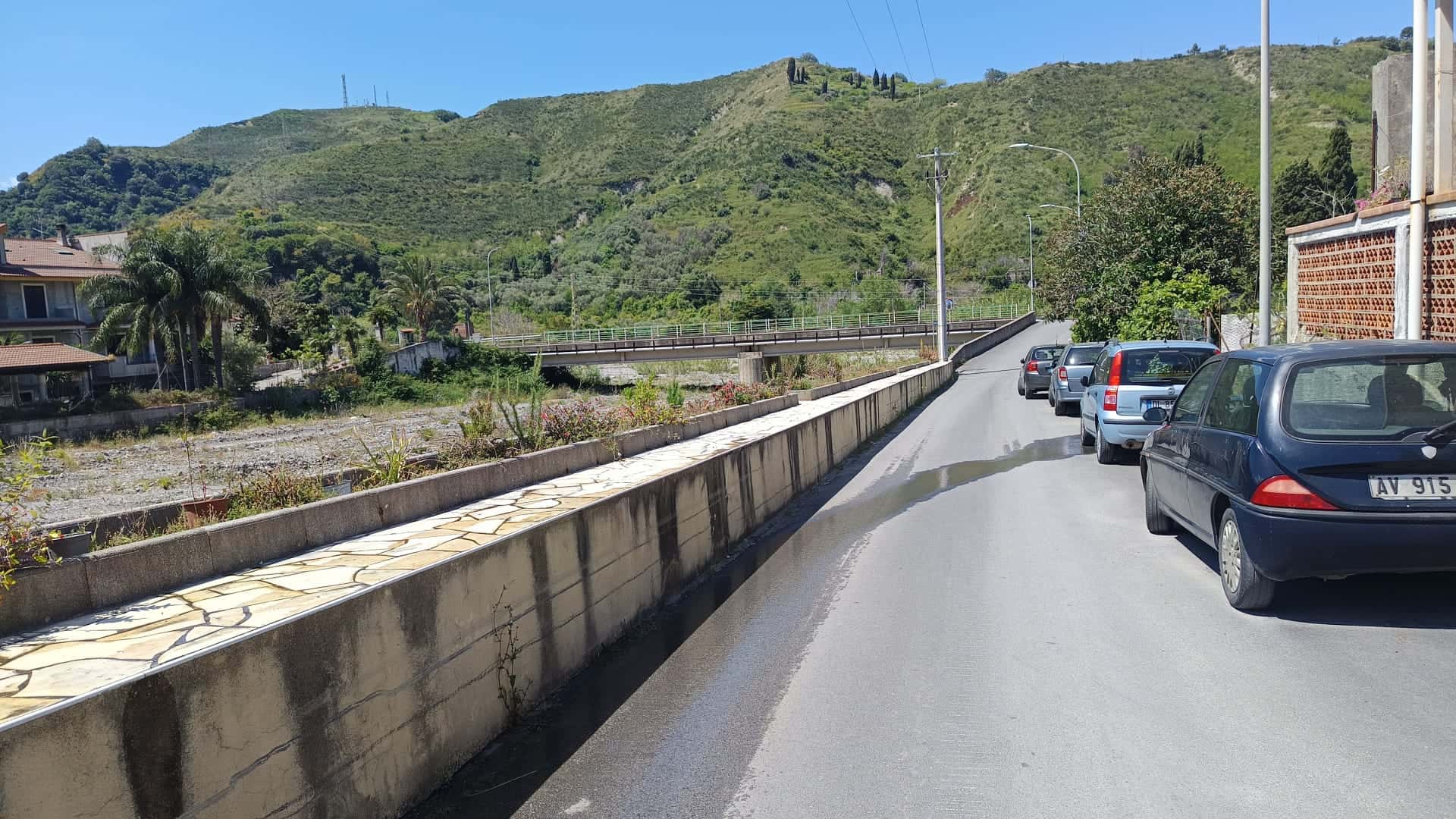 Ampia strada di montagna con auto parcheggiate, sfondo di colline verdi e cielo sereno, vista panoramica di un'area residenziale in un contesto naturale e tranquillo.