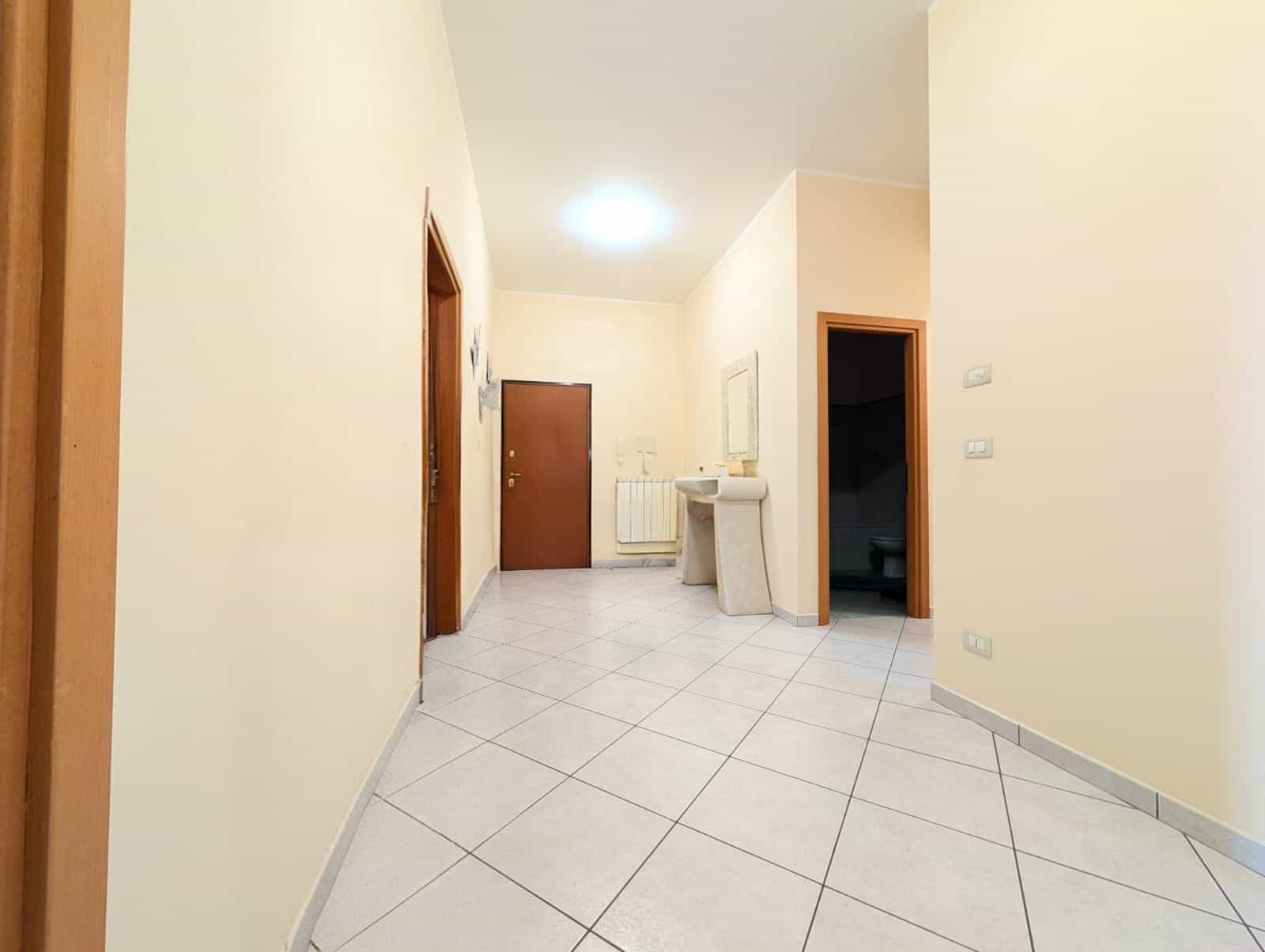 Ingresso di un appartamento con pareti beige, porta d'ingresso, radiatore e lavabo, pavimento in piastrelle chiare, ambiente luminoso e accogliente.