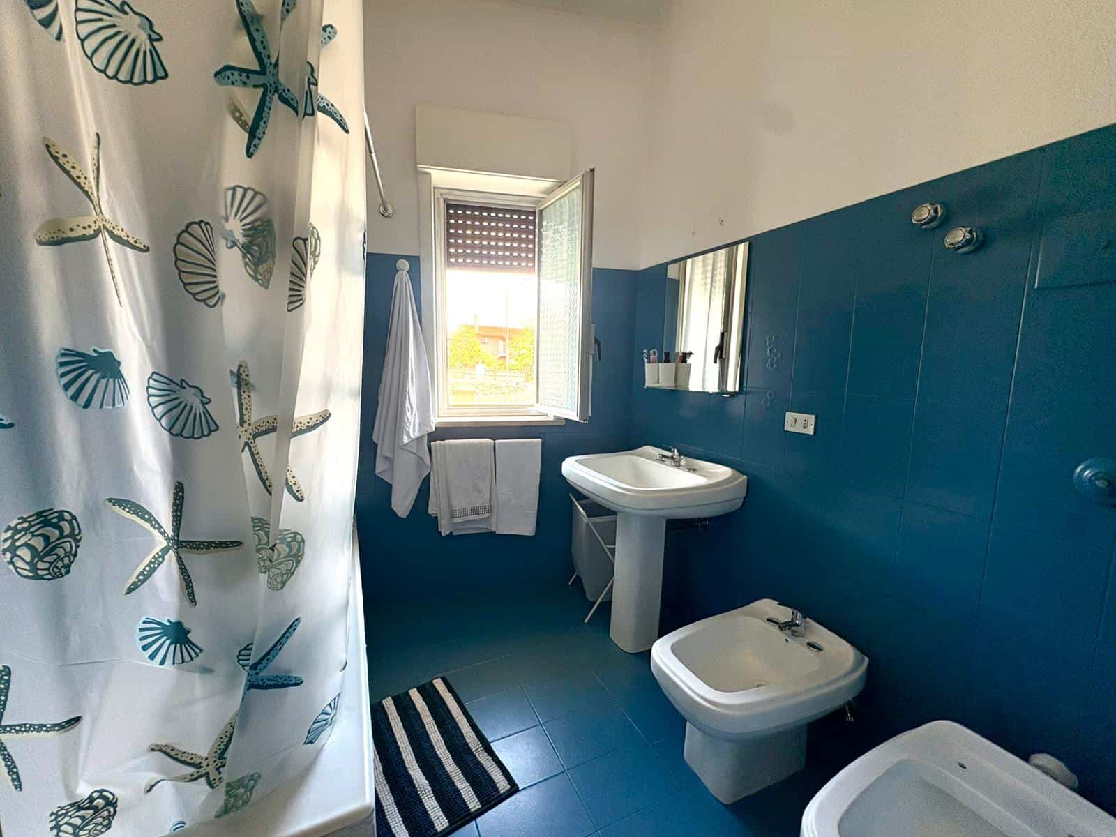 Ampia stanza da bagno con lavabo, bidet e doccia, illuminata da luce naturale proveniente da una finestra aperta, con pareti blu e accessori essenziali per il comfort quotidiano.