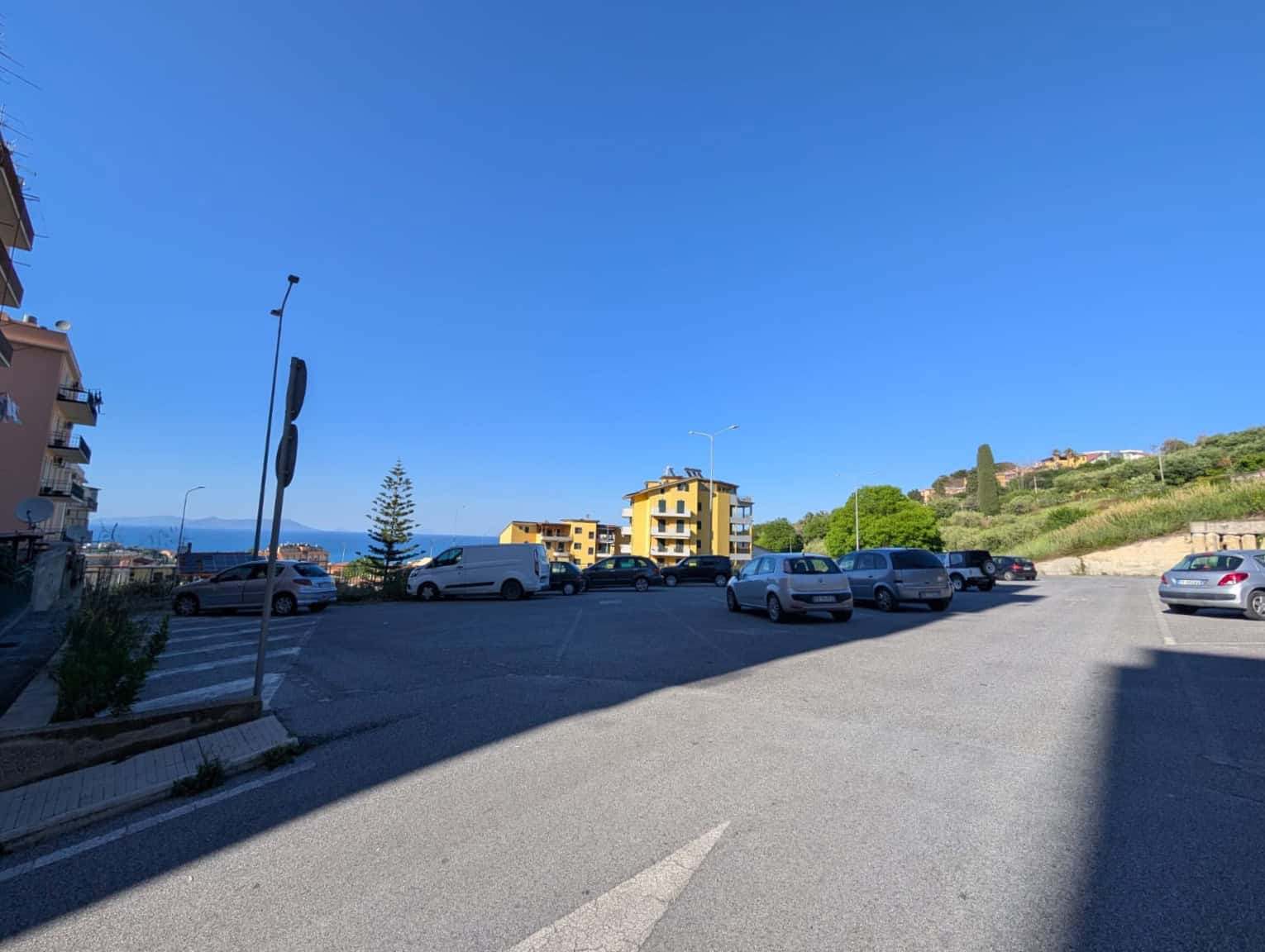 Auto in sosta in un parcheggio all'aperto con vista sul mare e sul paesaggio circostante, sotto un cielo sereno e azzurro, con edifici residenziali e vegetazione verde.